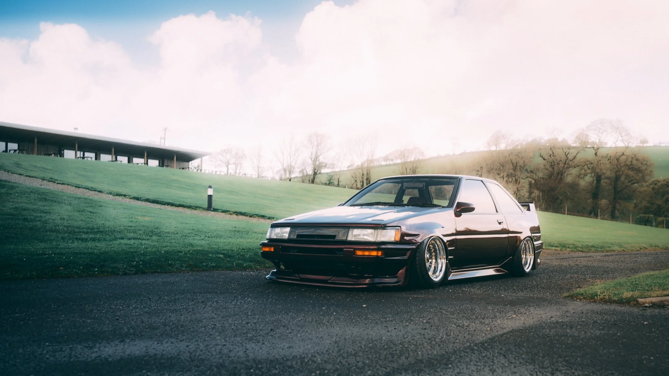 A black vintage sports car with lowered stance and aftermarket wheels parked on a paved surface in front of a grassy hill with a modern building and trees in the background under a partly cloudy sky.