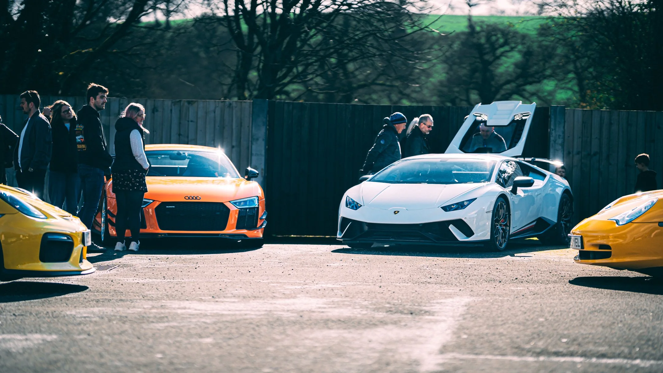 People standing around luxury sports cars, including a white Lamborghini and an orange Audi, at an outdoor car gathering on a paved lot with a wooden fence and trees in the background.