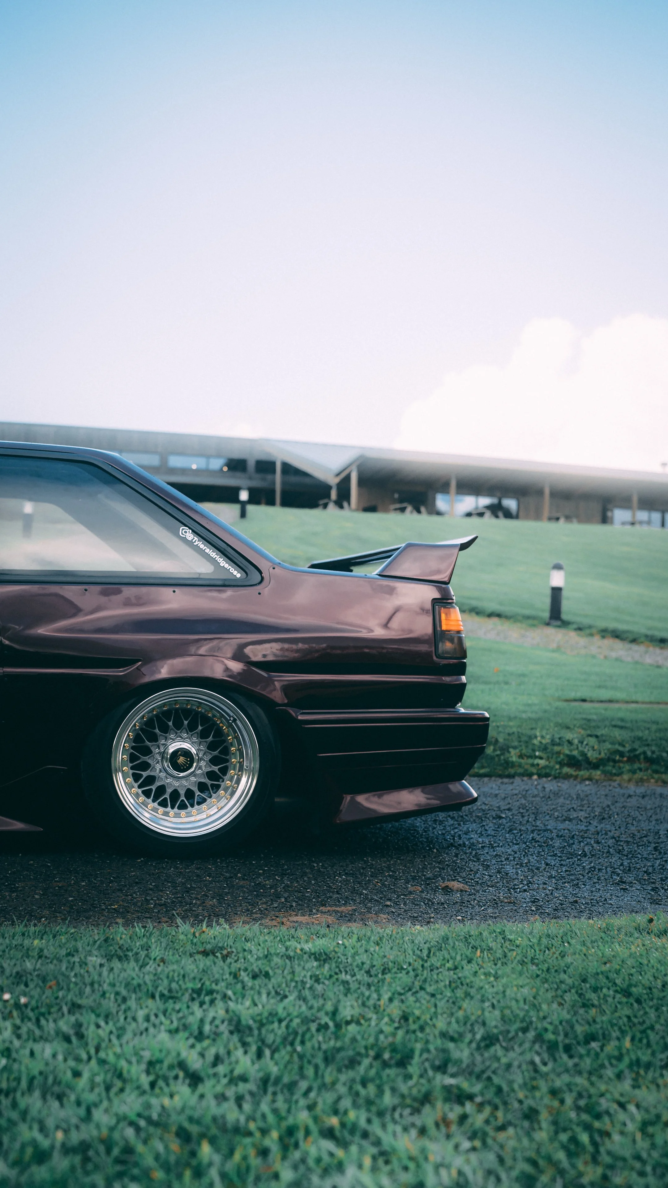 Close-up of a vintage maroon car's rear on a grassy area with modern building in background.
