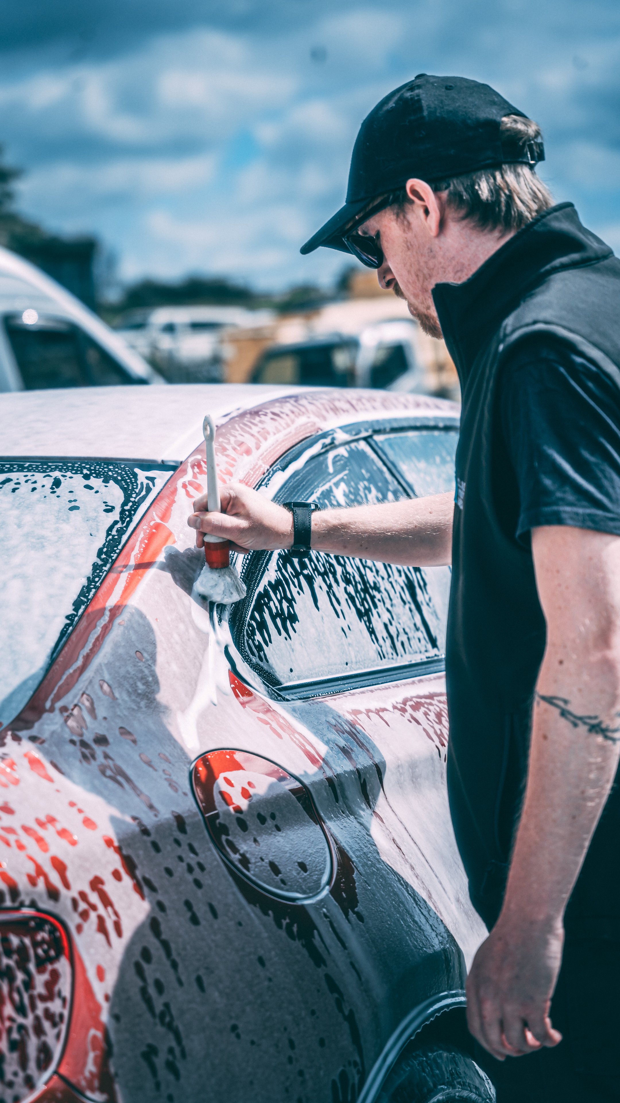 Person in black shirt and sunglasses using a brush to wash a red and black car at a car wash facility.