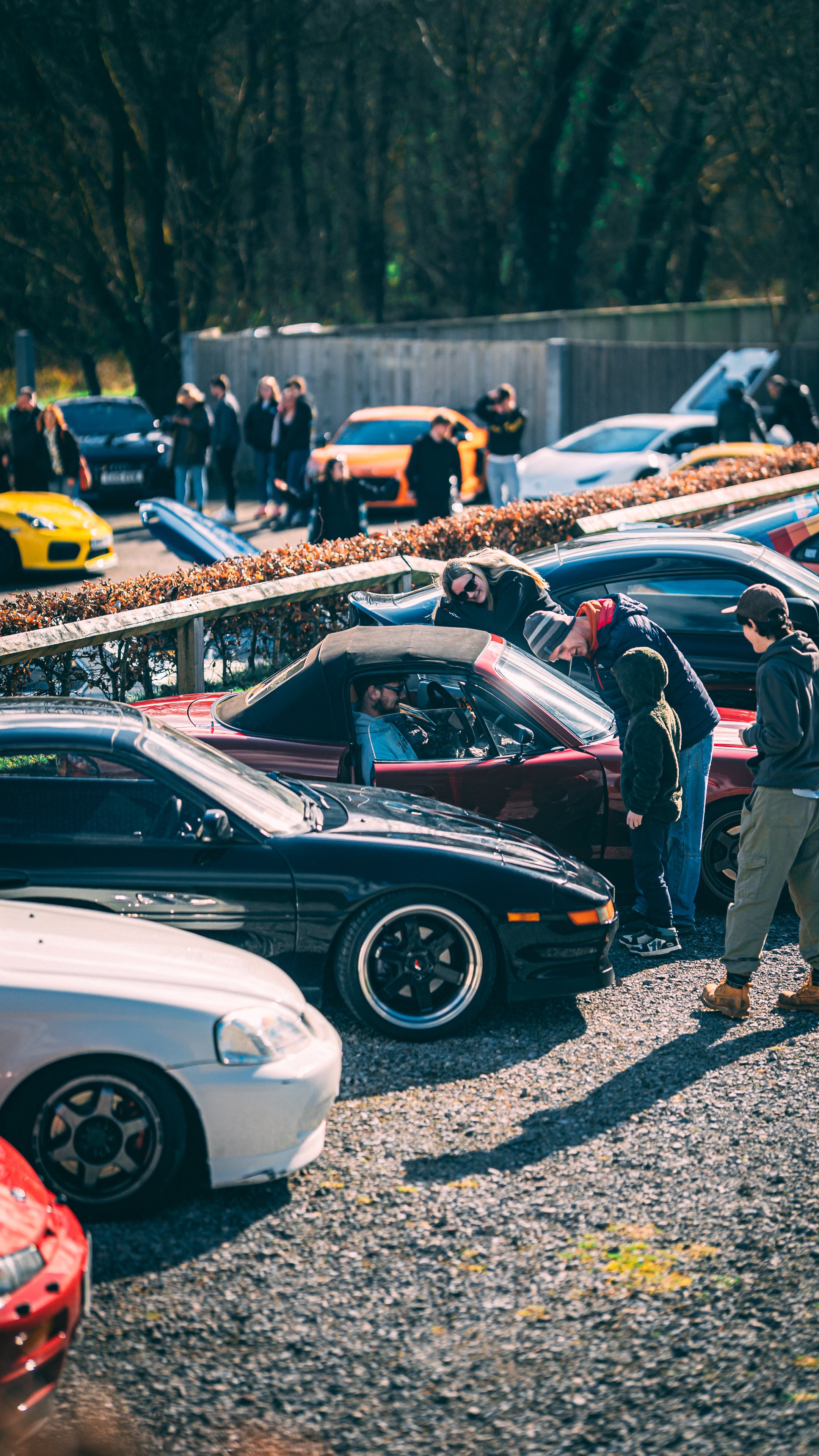 People gathered around a red sports car, engaged in conversation, with other luxury cars parked nearby at a car meet event, outdoors on a sunny day.