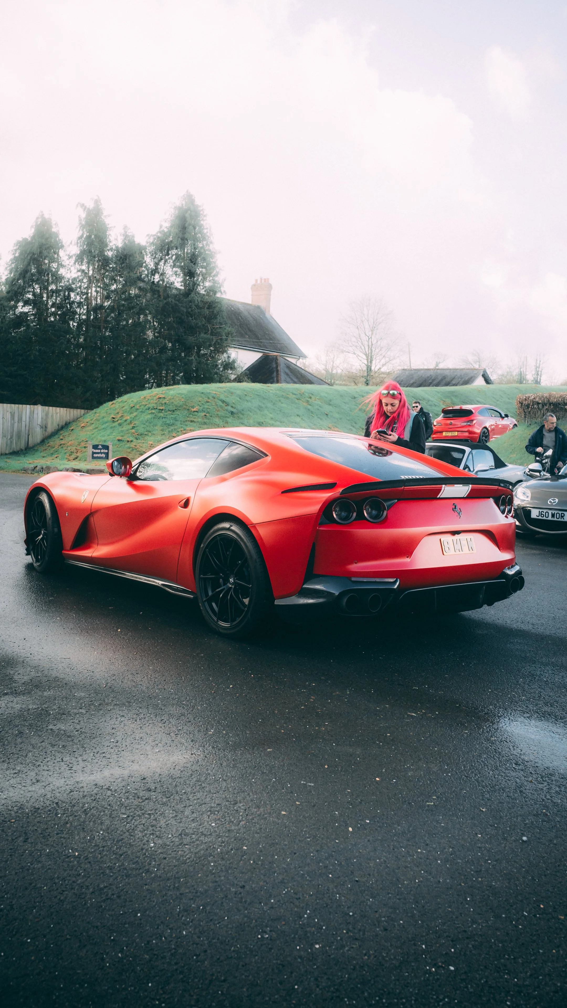 Red Ferrari sports car parked on a wet road, with a woman with pink hair and sunglasses looking at her phone in the background.