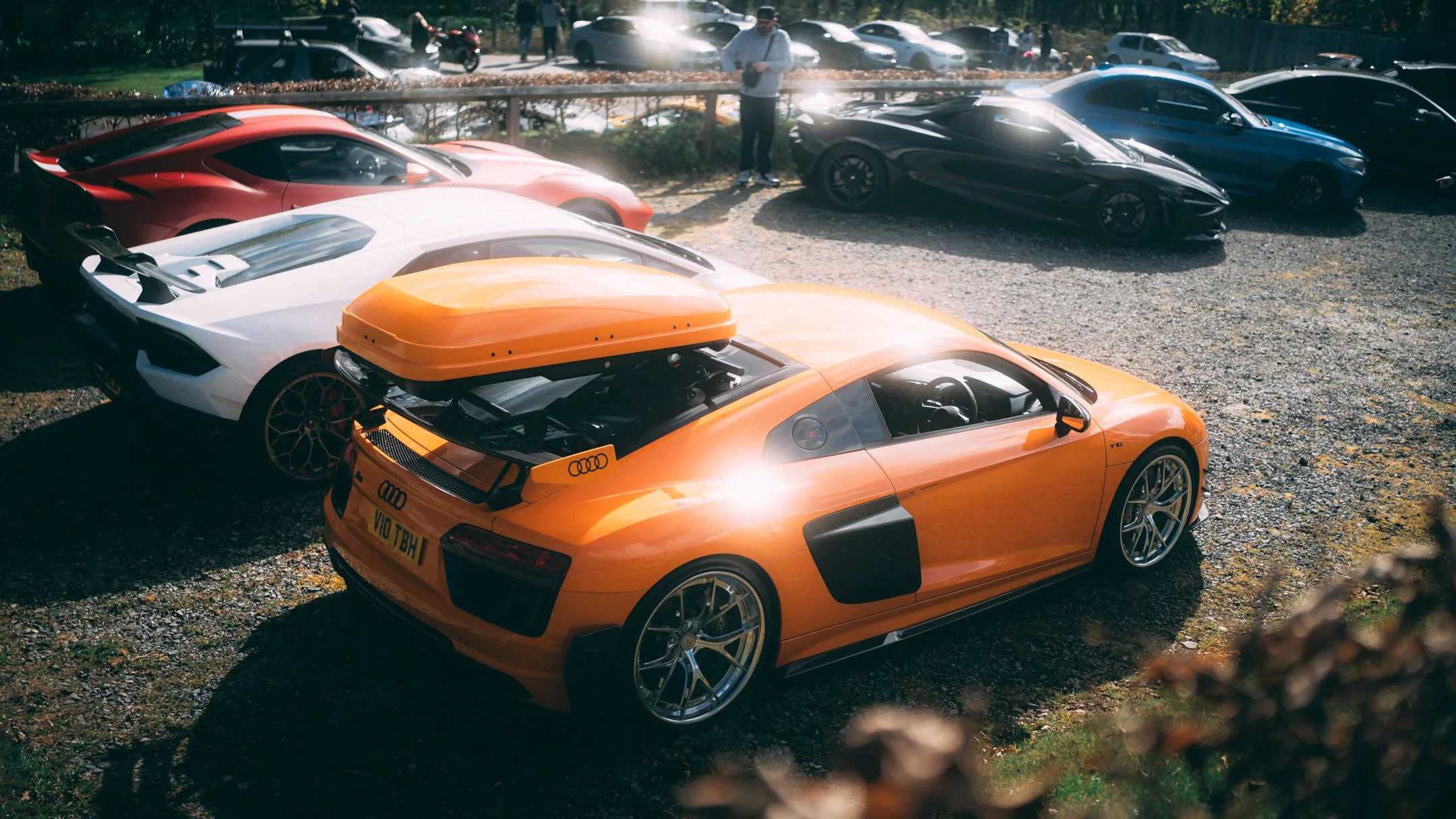 Multiple high-end sports cars parked outdoors in a gravel area with a few people walking and standing nearby, trees and other vehicles visible in the background.