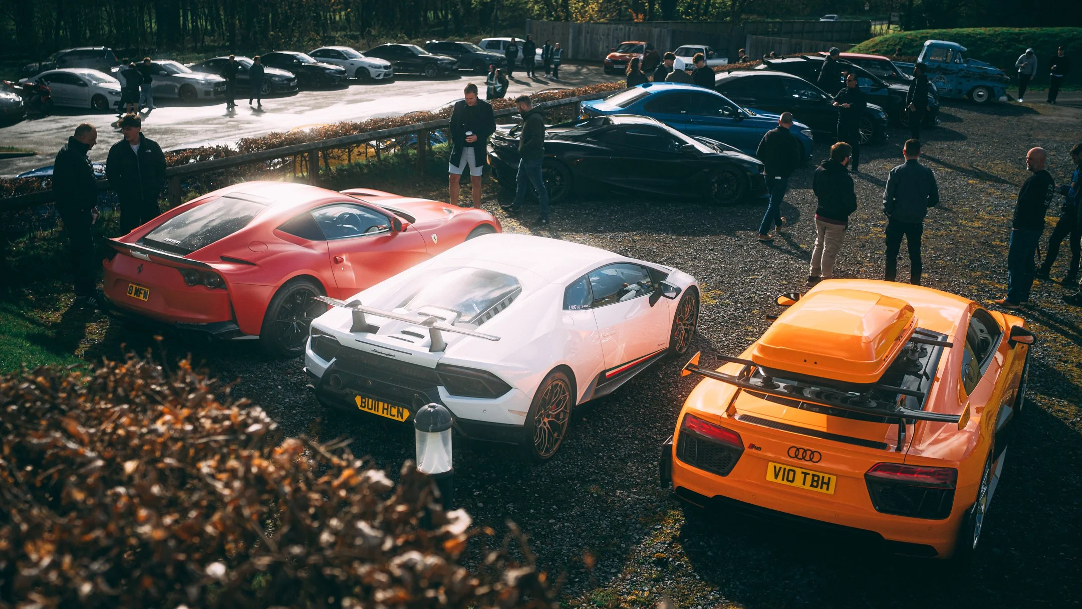 A parking lot filled with luxury sports cars, including a red Ferrari, a white Lamborghini, and an orange Audi R8, with people standing around and conversing on a sunny day.
