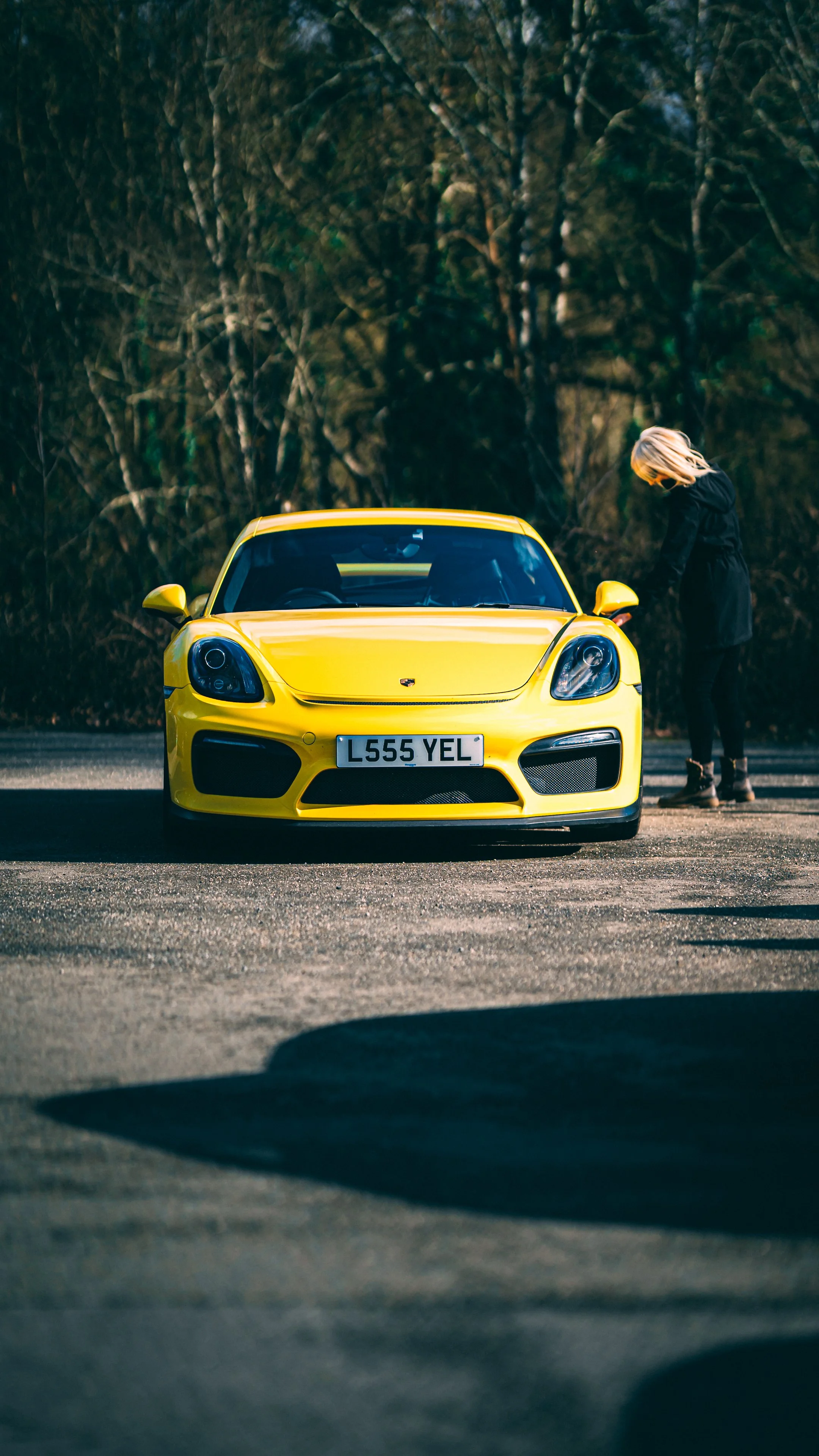 A yellow sports car parked on a road with a woman in a black jacket standing beside it, looking down. There are trees and foliage in the background.