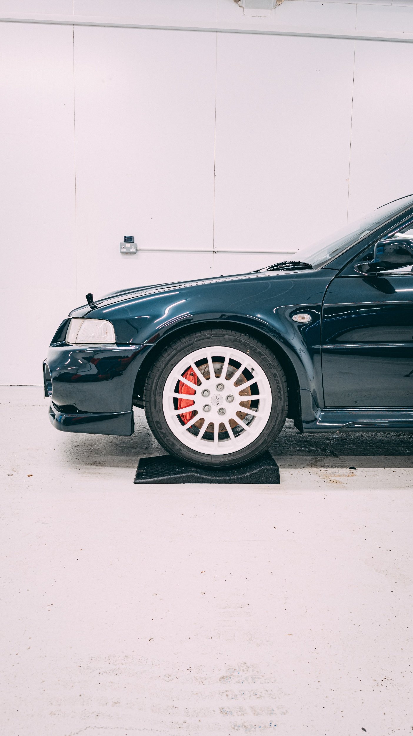 Black sports car with white racing wheels and red brake calipers, parked indoors on a black wheel chock against a white wall.