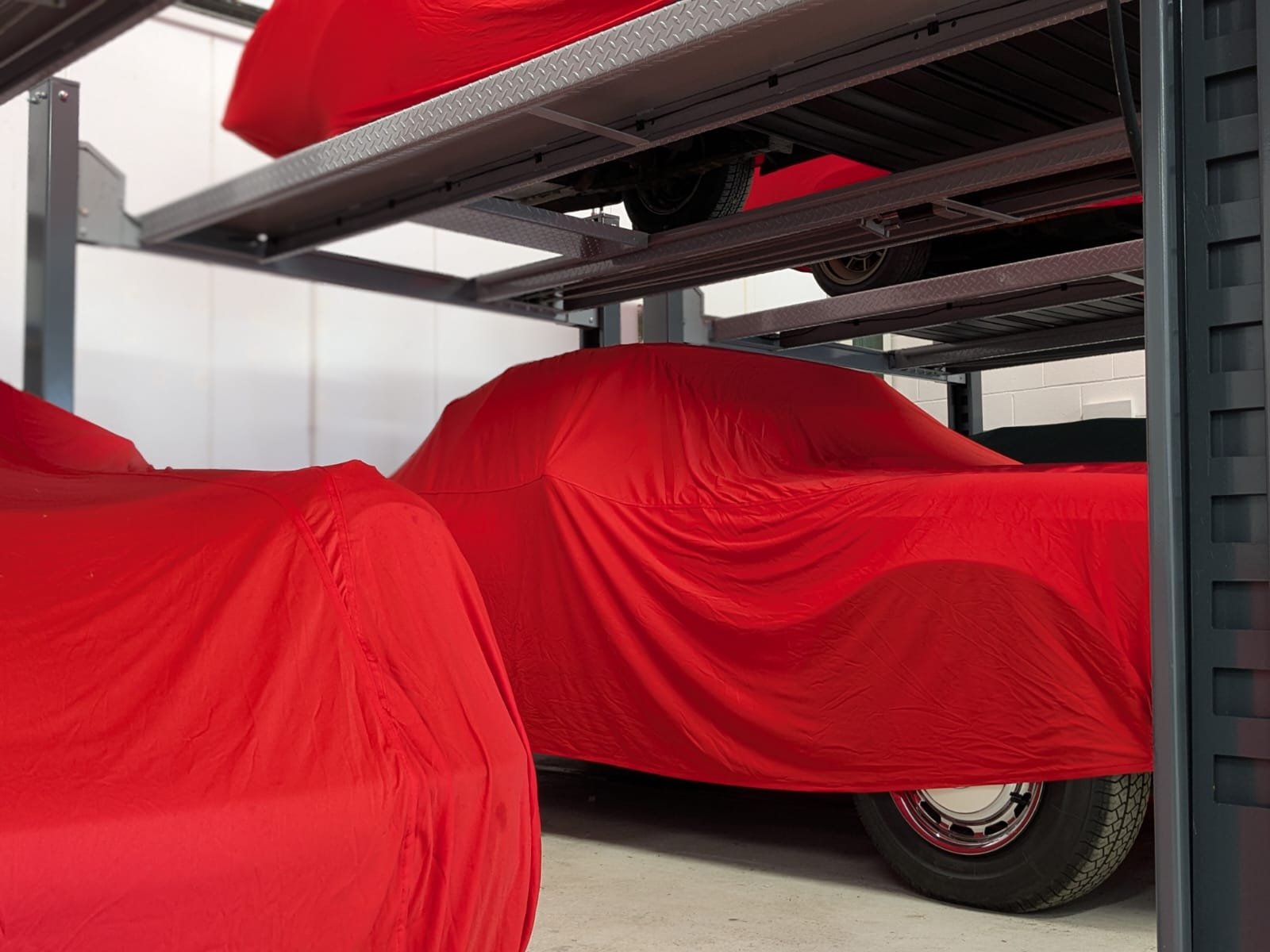 Three cars covered with red protective covers, stored on a metal car rack in a garage.