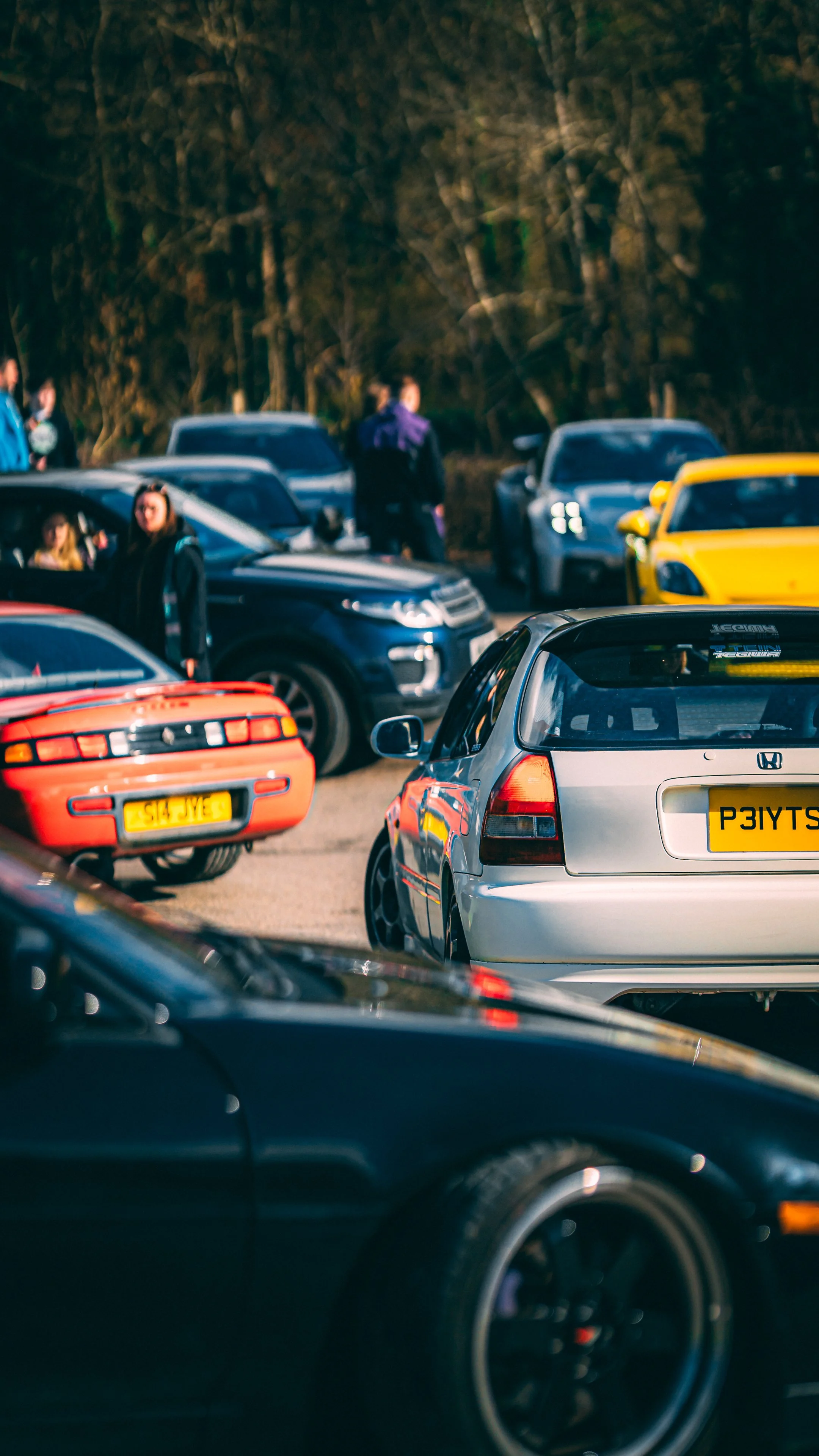 A parking lot filled with various parked cars, including a red sports car, a silver hatchback, and a yellow car, with some people standing and walking near the vehicles and trees in the background.