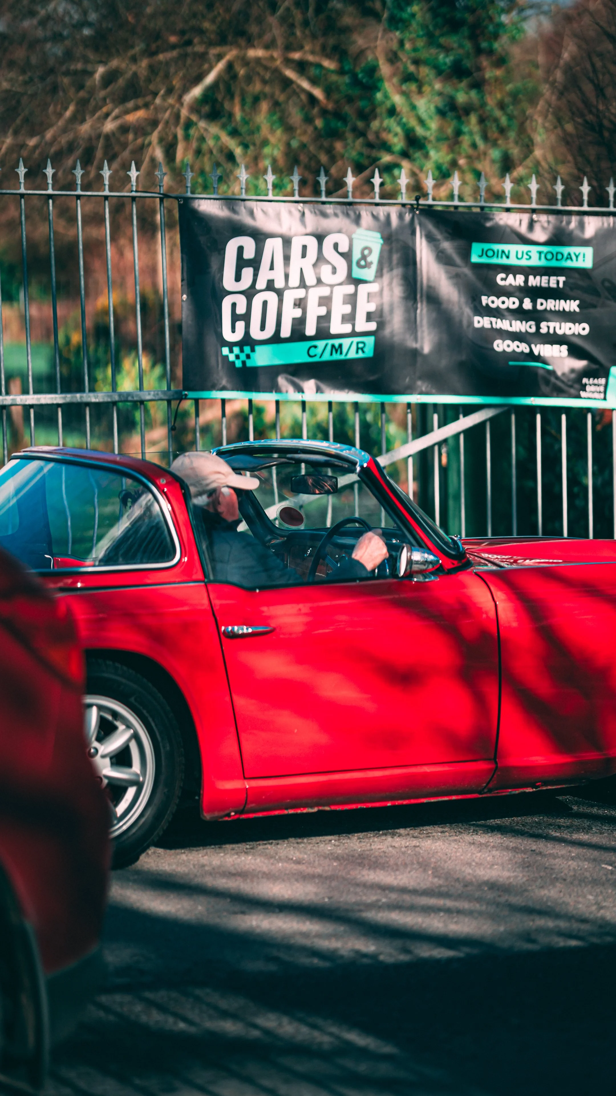 Red vintage car parked with a driver inside, in front of a black fence with a promotional banner advertising a car meet event that includes food and drinks.