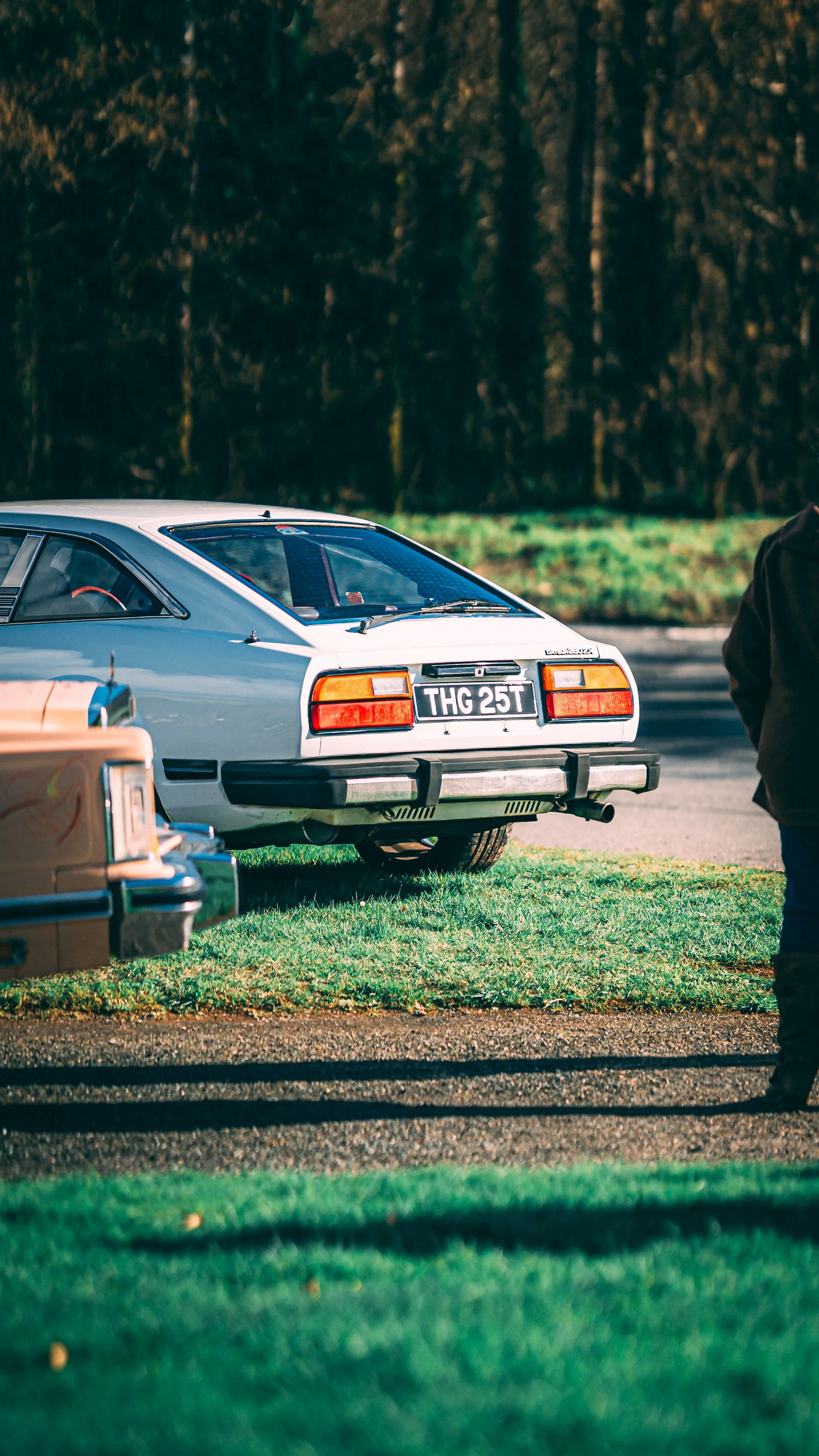 A silver vintage car parked on the grass next to a gravel driveway with a partially visible person standing on the right side.