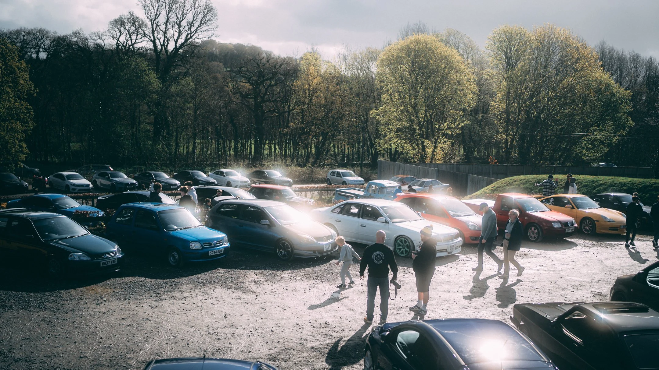 People walking and standing near a parking lot filled with cars, with trees in the background under a cloudy sky.