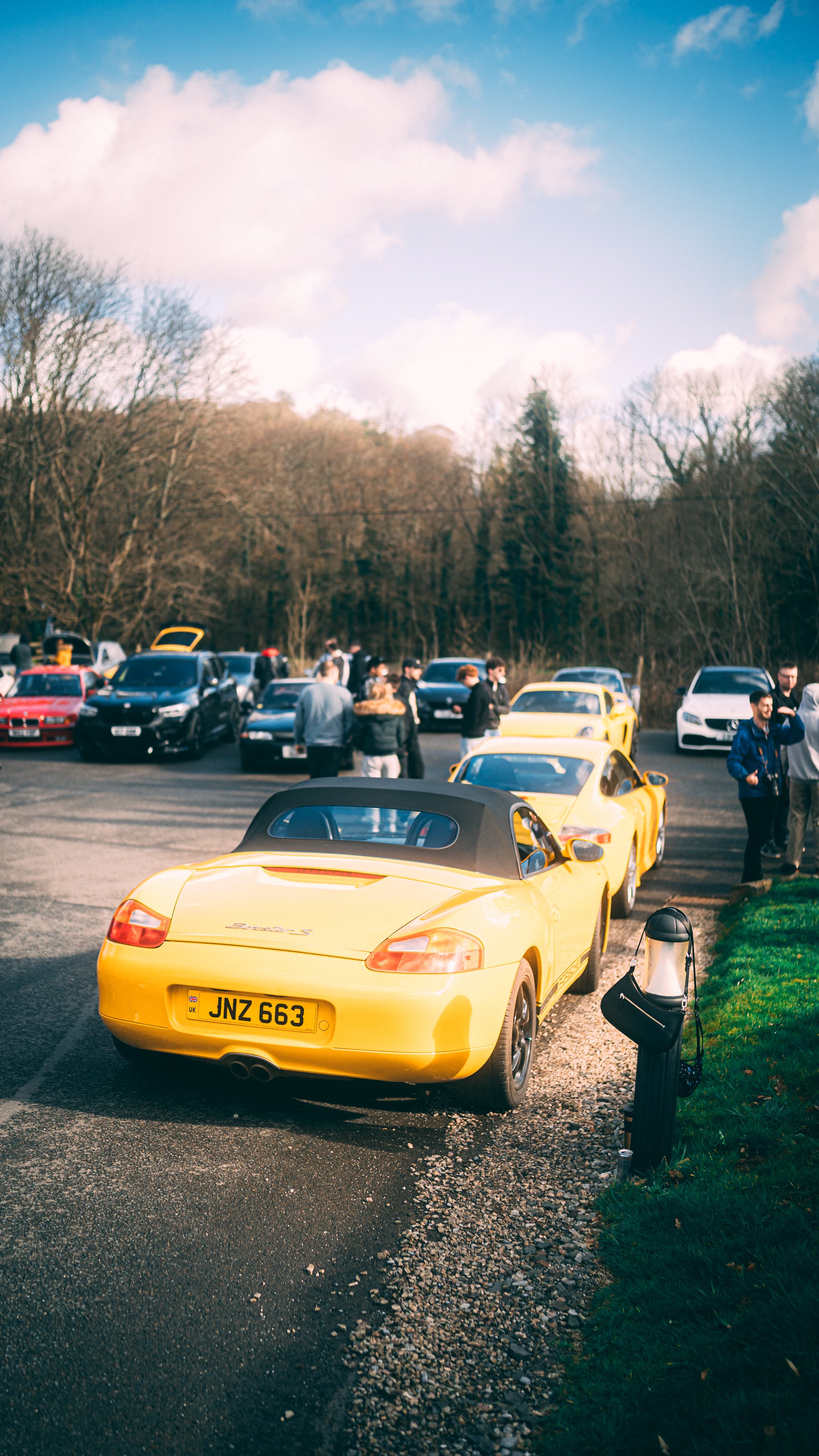 A yellow Porsche convertible parked in a lot filled with various cars and people gathered around, with trees and a partly cloudy sky in the background.