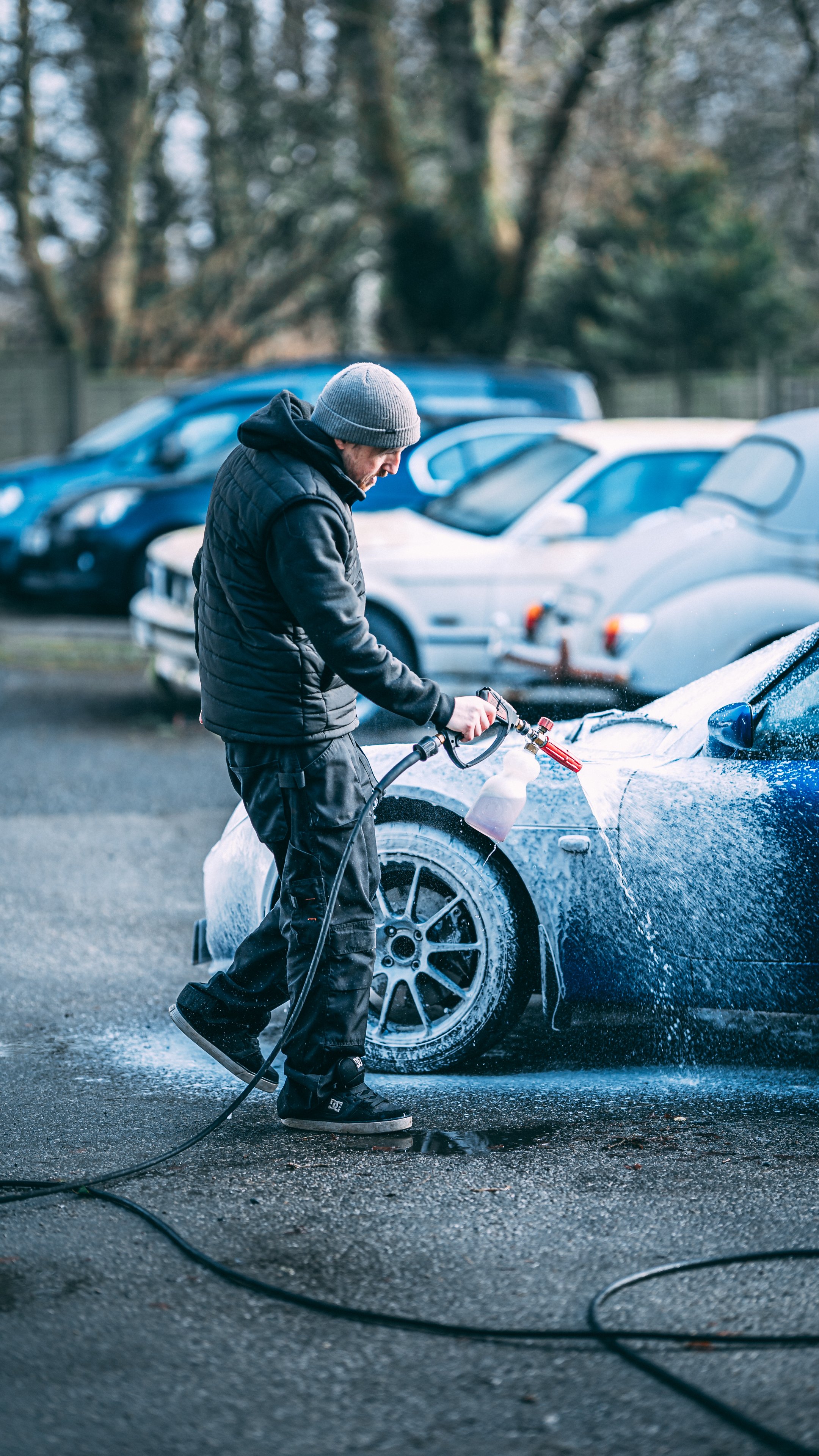 A man wearing a gray beanie and black jacket pressure washing a sports car with foam, in a parking lot with several cars and trees in the background.