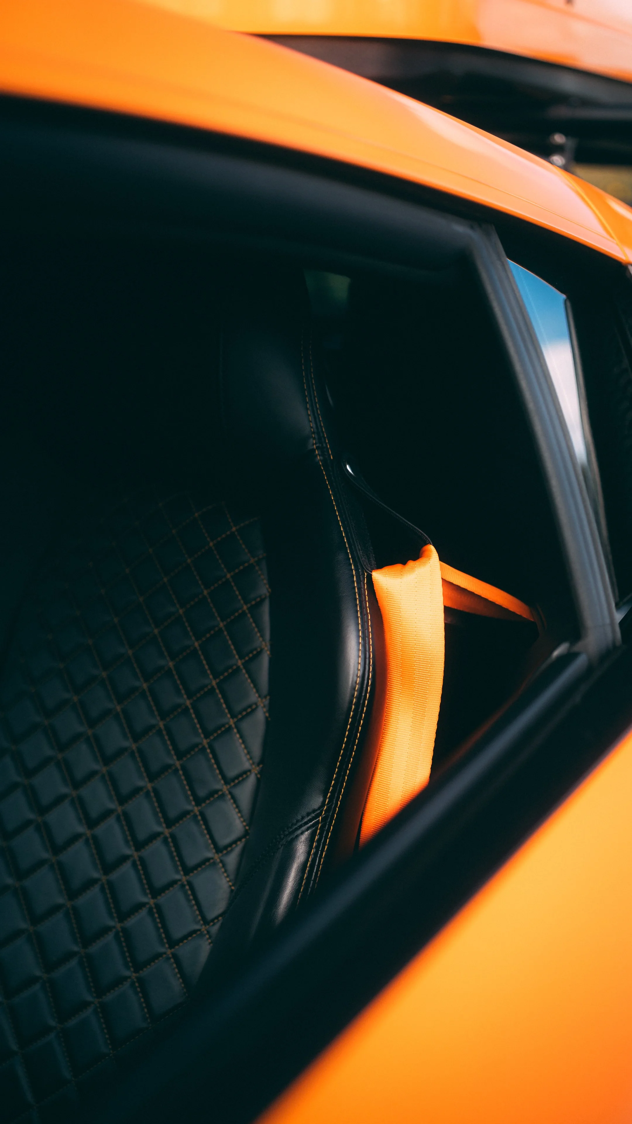 Close-up of the black interior seat with orange stitching and a bright orange headrest strap in a sports car.