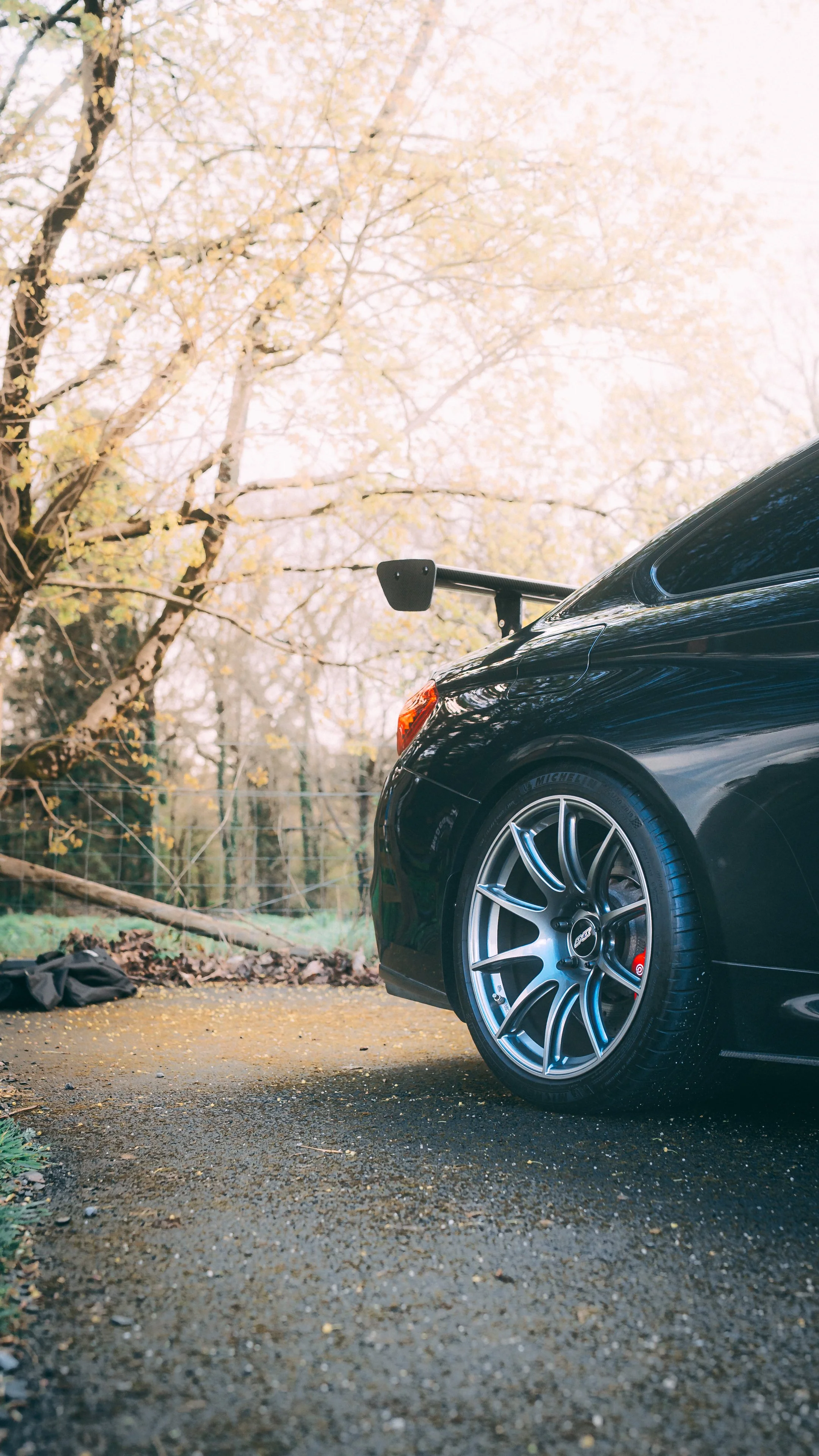 The front part of a black sports car with a large rear wing parked on an autumn day with trees in the background.