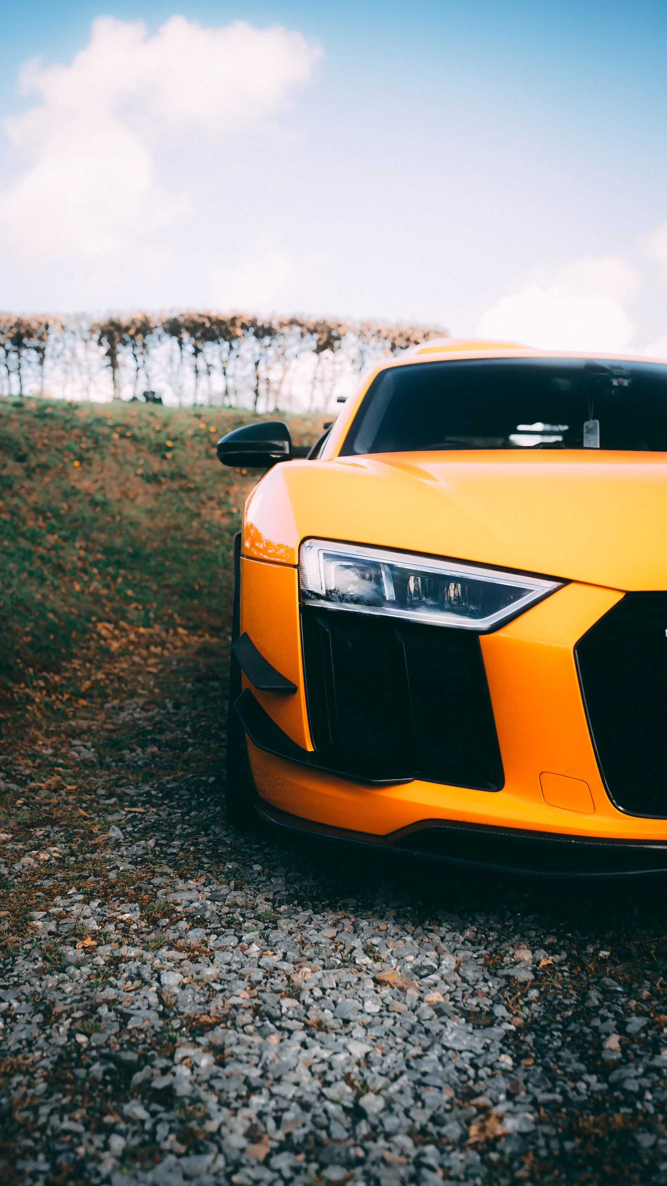 Close-up of the front of a yellow sports car parked on a gravel path with a background of trees and a partly cloudy sky.