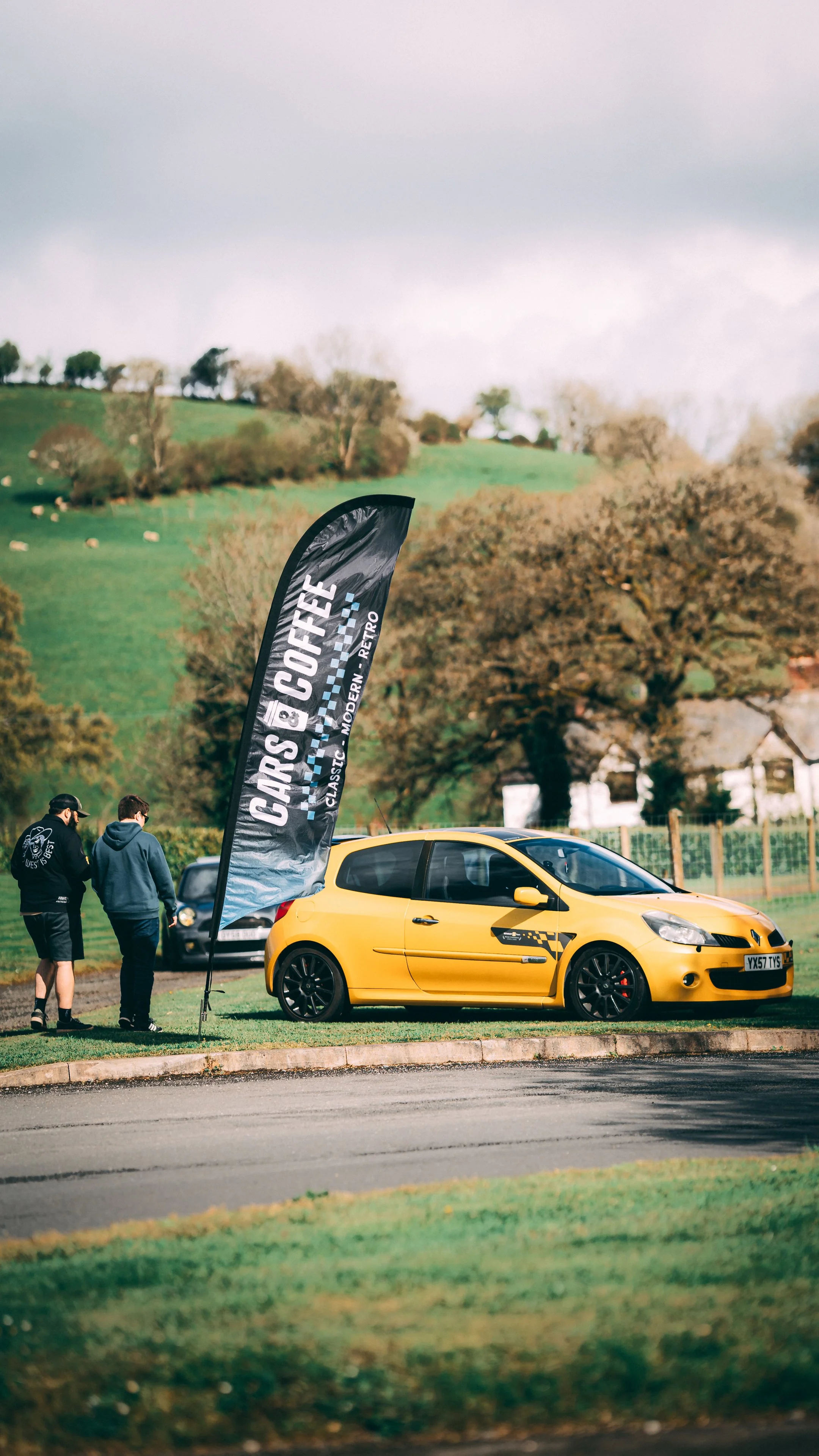 Yellow sports car parked on grass with a black banner reading 'CARS & COFFEE, Classic, Modern, Retro'. Two men walking nearby, in a rural setting with trees and rolling hills in the background.