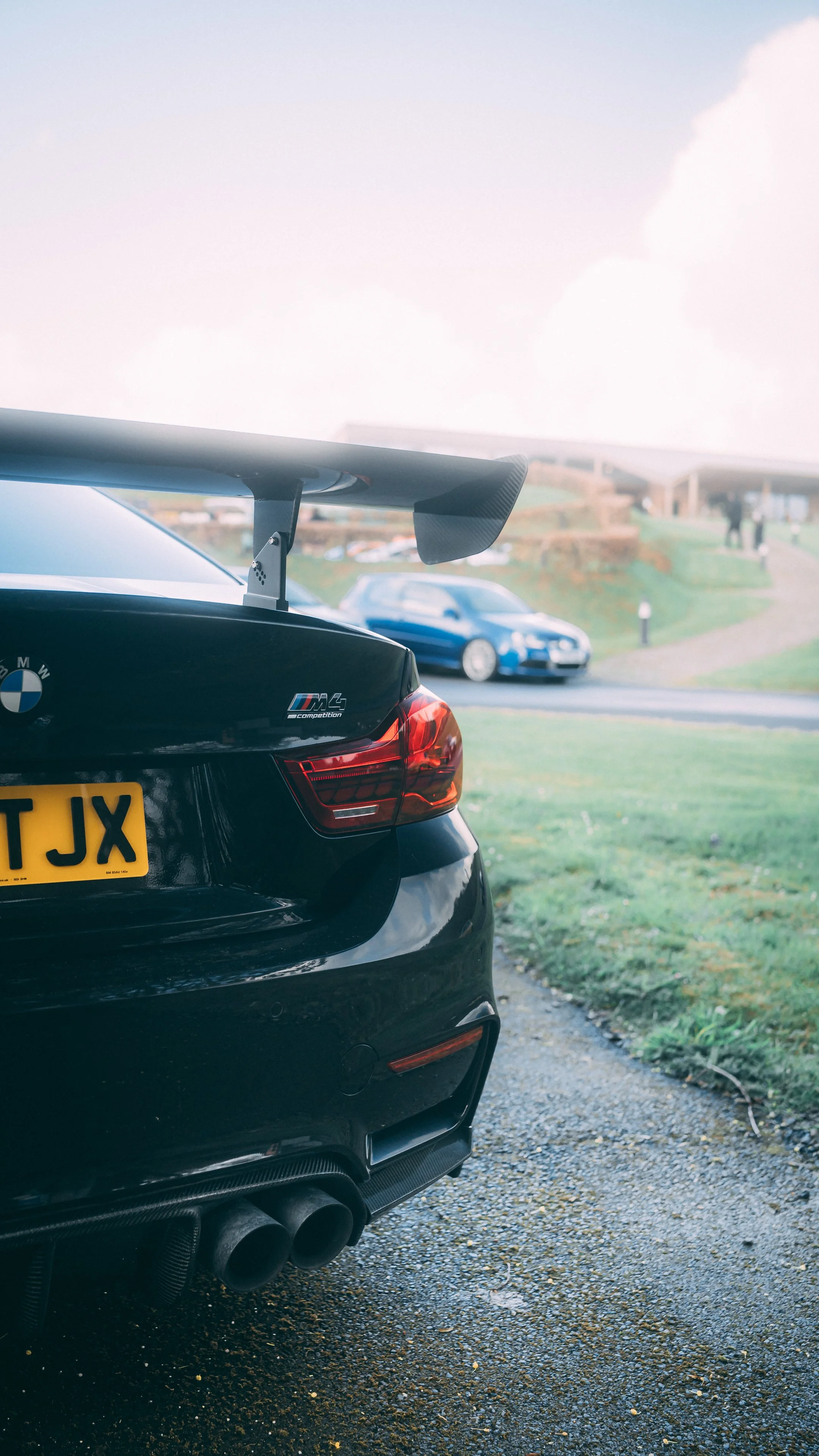 Close-up of the rear of a black BMW M4 car with an M Performance badge and a large rear wing, parked on a gravel driveway with grassy areas and other cars in the background.
