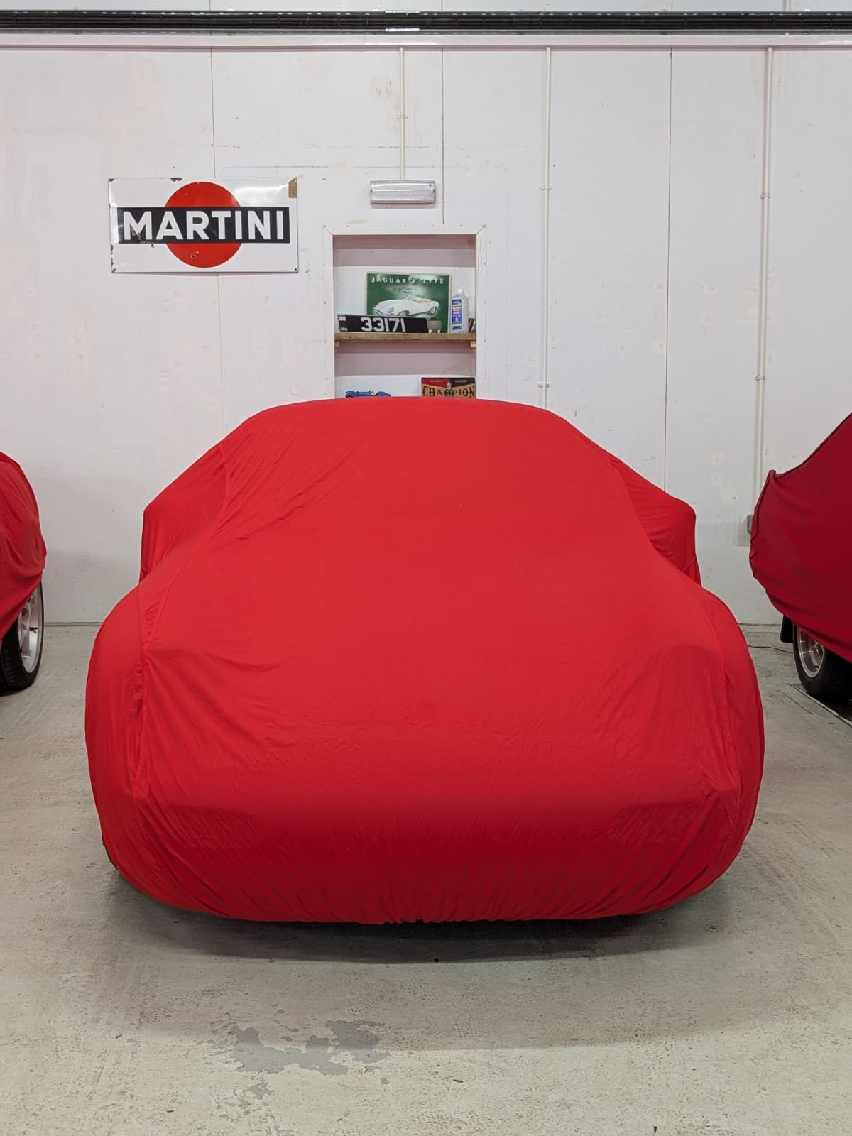 A car covered with a red car cover in a garage, with a white wall and a shelf with car-related items and a vintage sign in the background.