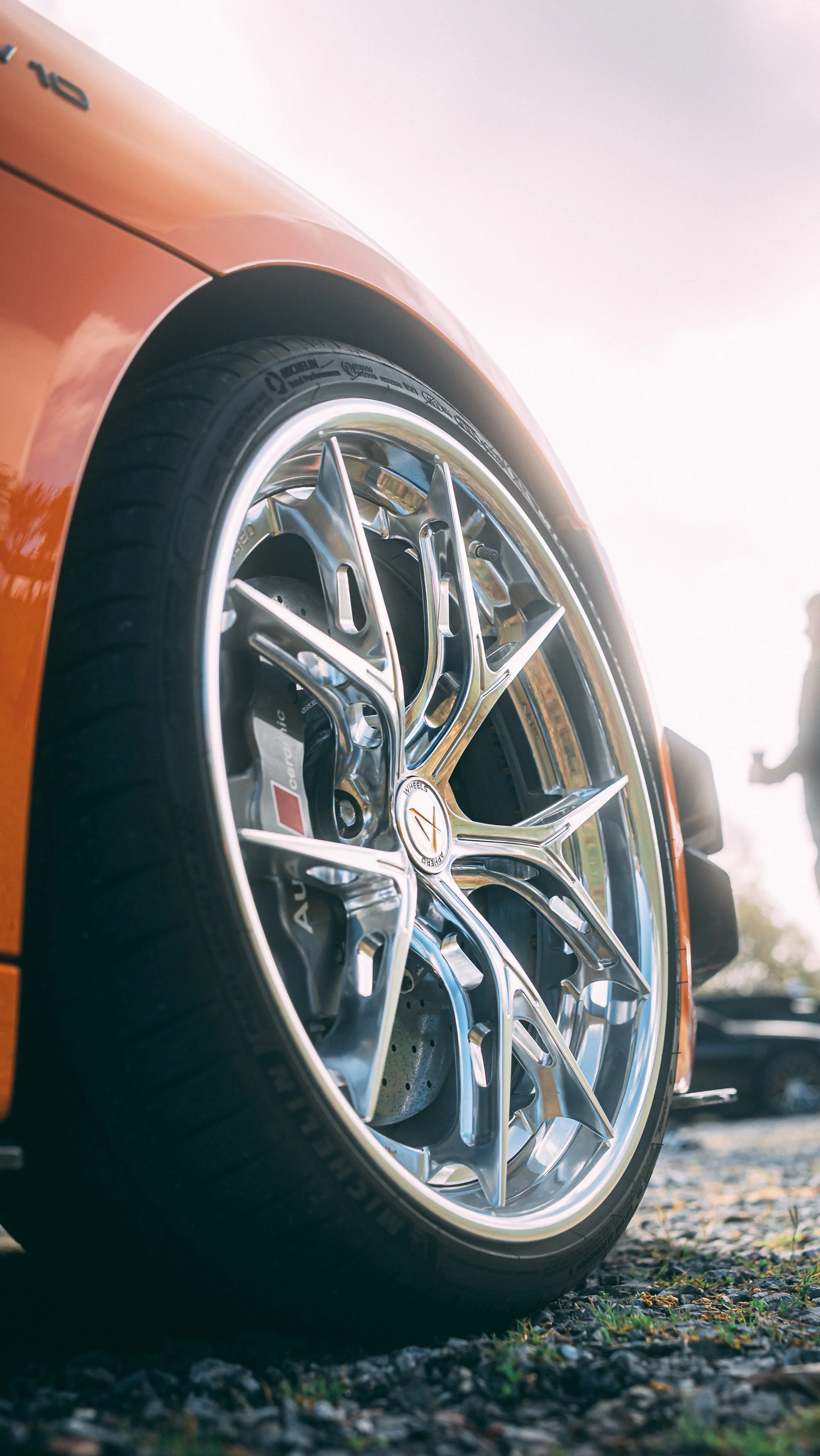 Close-up of a sleek orange sports car wheel with a shiny metallic rim and a black tire on a gravel surface, with a blurred background.