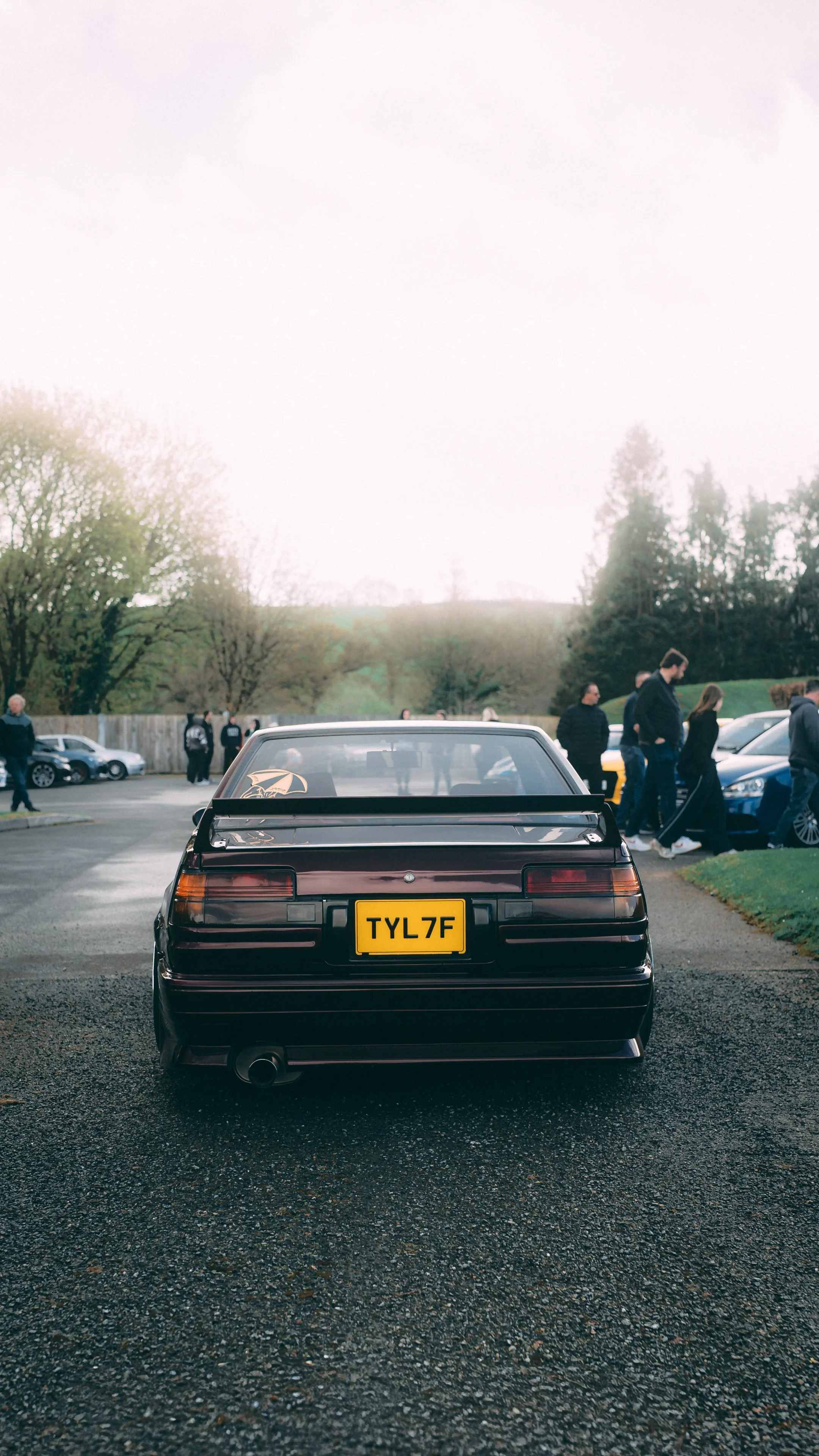 A rear view of a vintage black car with a yellow license plate reading 'TYL7F', parked in a lot filled with people and other cars, with trees and a cloudy sky in the background.