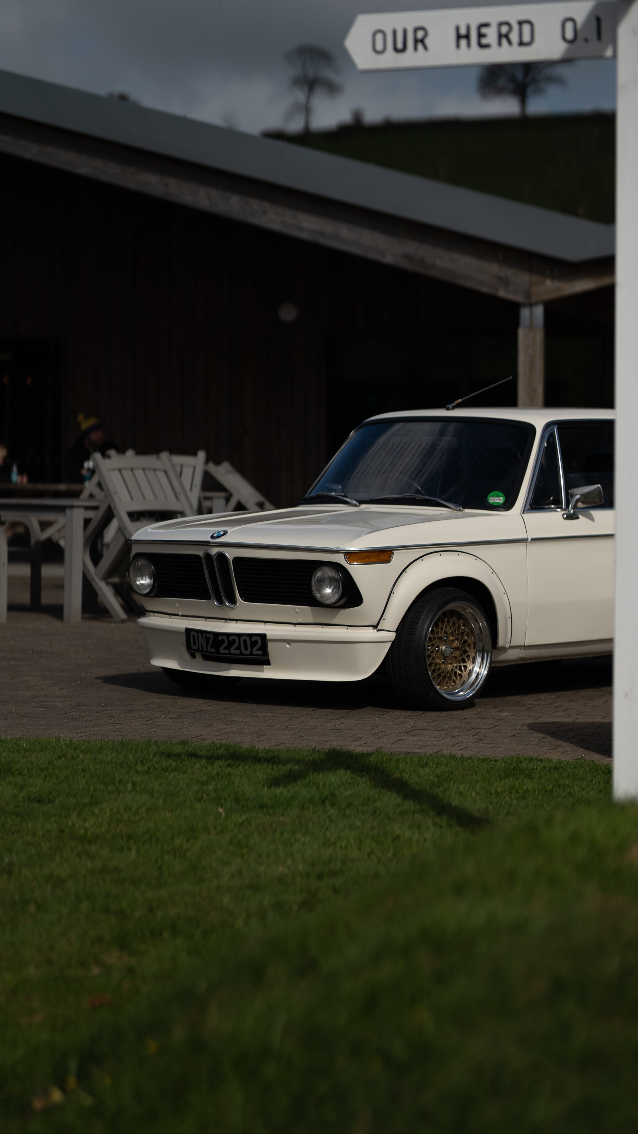 A white vintage BMW car parked on a paved area, with gold alloy wheels and a black grille, in front of a dark building. A sign overhead reads "OUR HERD 0.1" and there are trees on a hill in the background.