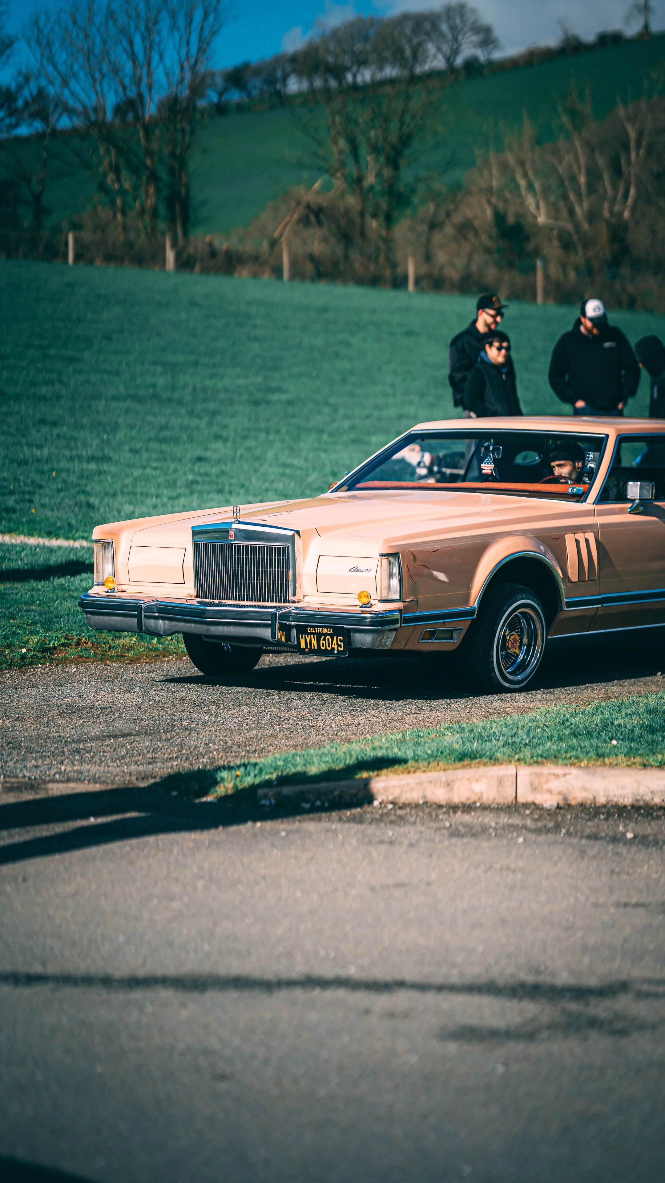 A vintage beige Cadillac parked on a roadside with three people standing in the background on a grassy area. The driver, wearing a cap, is inside the car. The background features green fields, trees, and a partly cloudy sky.