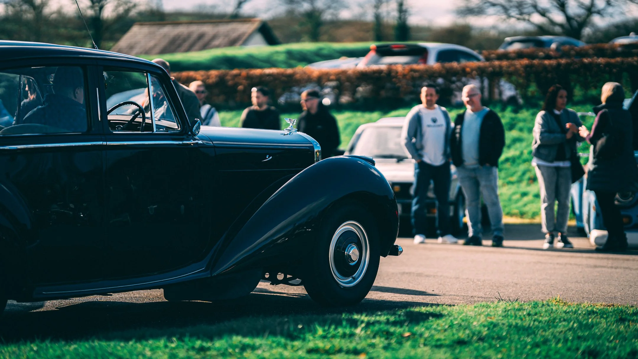 A group of people standing outdoors near a vintage black car, with trees and other vehicles in the background.