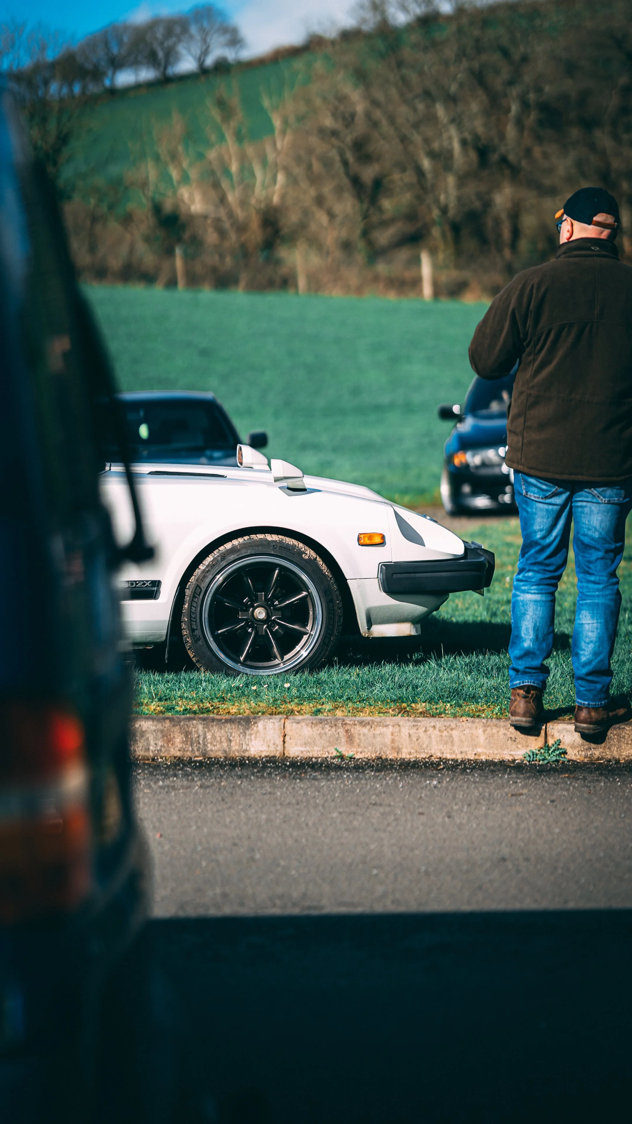 A man in a dark jacket, blue jeans, and a black cap stands next to a silver sports car parked on a grassy area. Other cars are visible in the background. The scene is outdoors with trees and rolling hills.