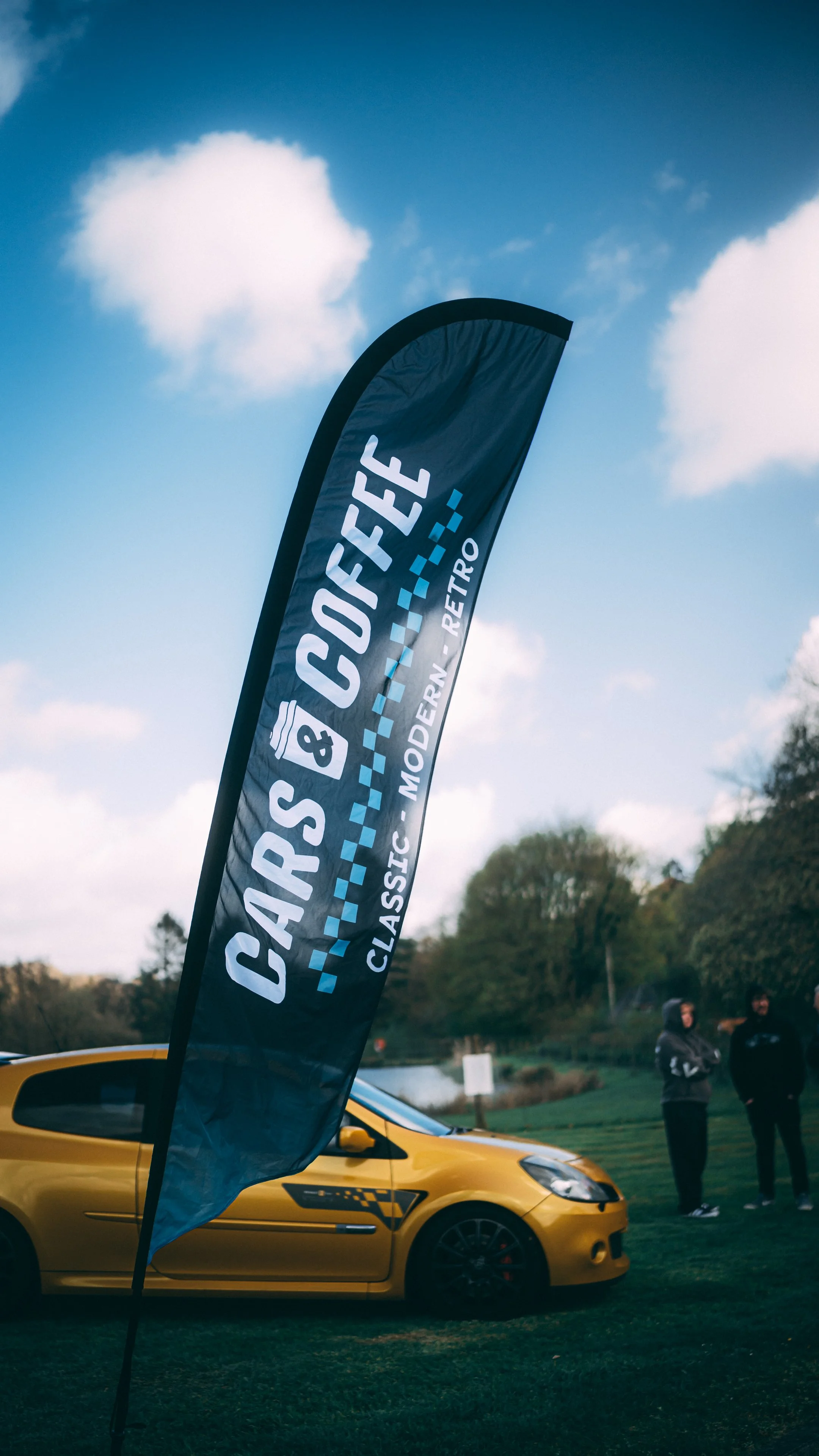 A yellow car parked outdoors next to a black flag with white and blue text that reads 'CARS & COFFEE CLASSIC MODERN RETRO'. There are three people standing in the background, and the scene is set on a grassy area with trees and a cloudy sky.