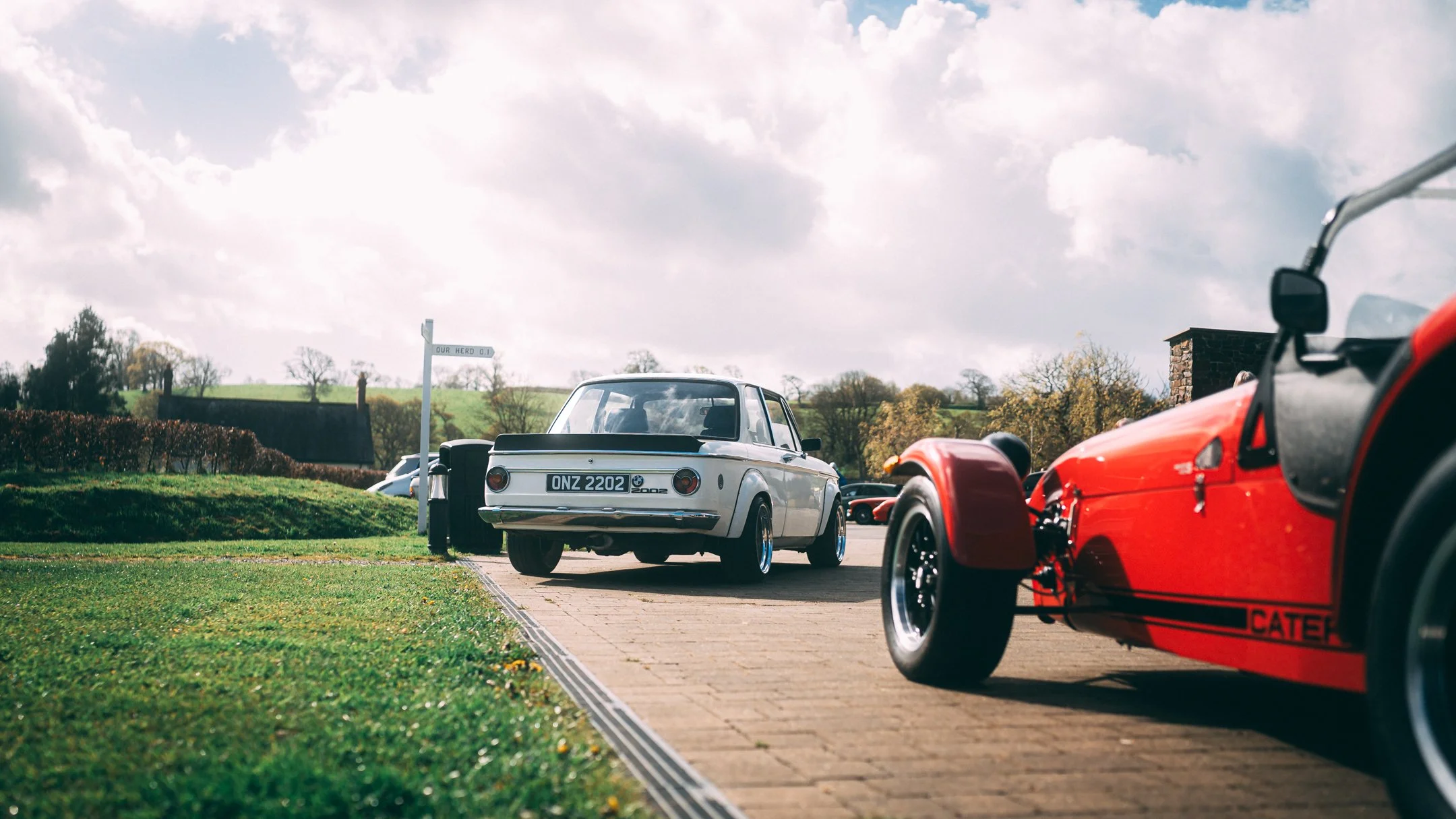A parking area with classic cars on display, including a white car in the center and a red sports car on the right, with a scenic landscape of green hills and trees under a partly cloudy sky in the background.