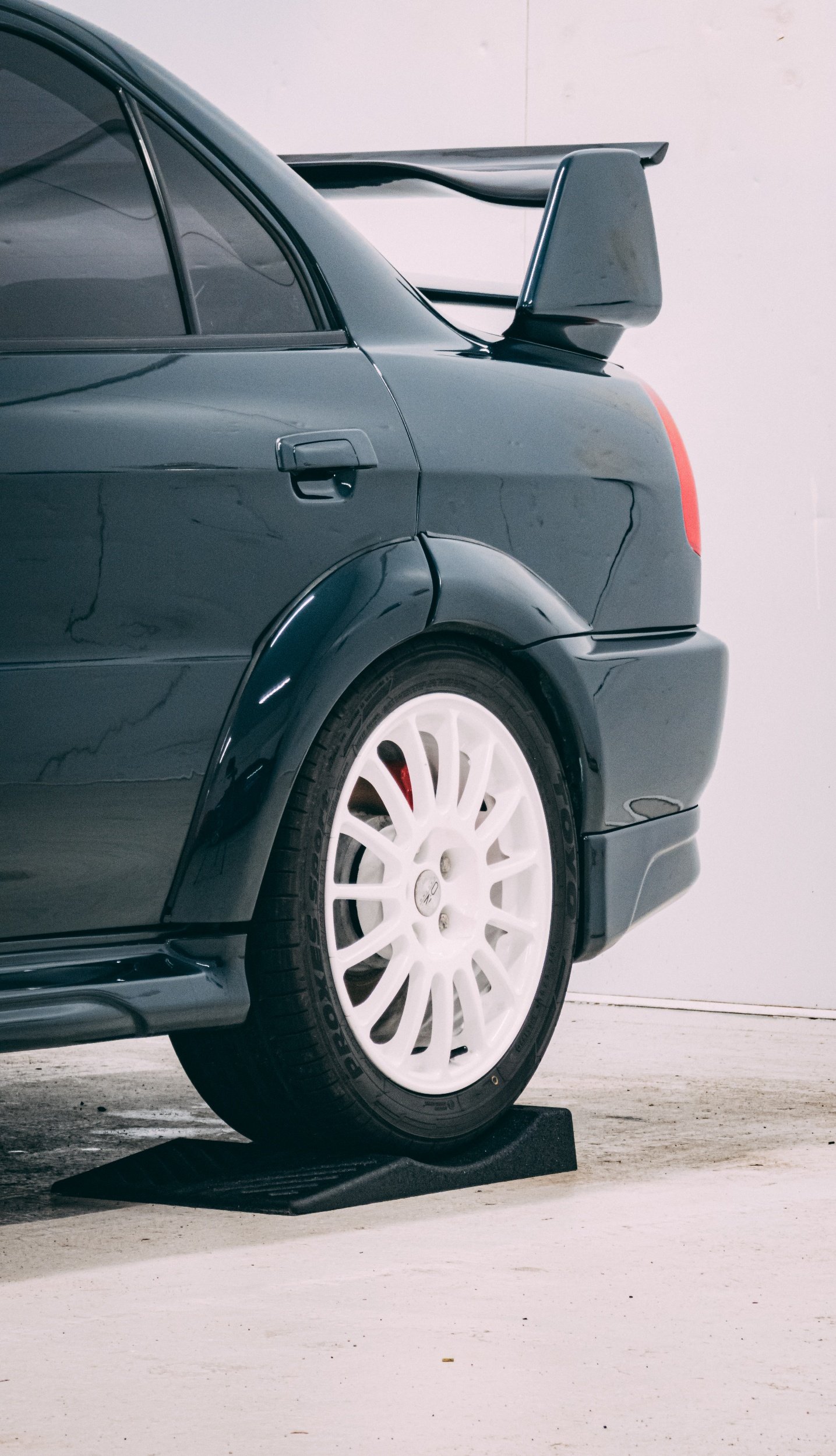 Close-up of the rear side of a black car with white racing wheels, parked inside a garage or studio, with a car wheel chock under the rear tire.