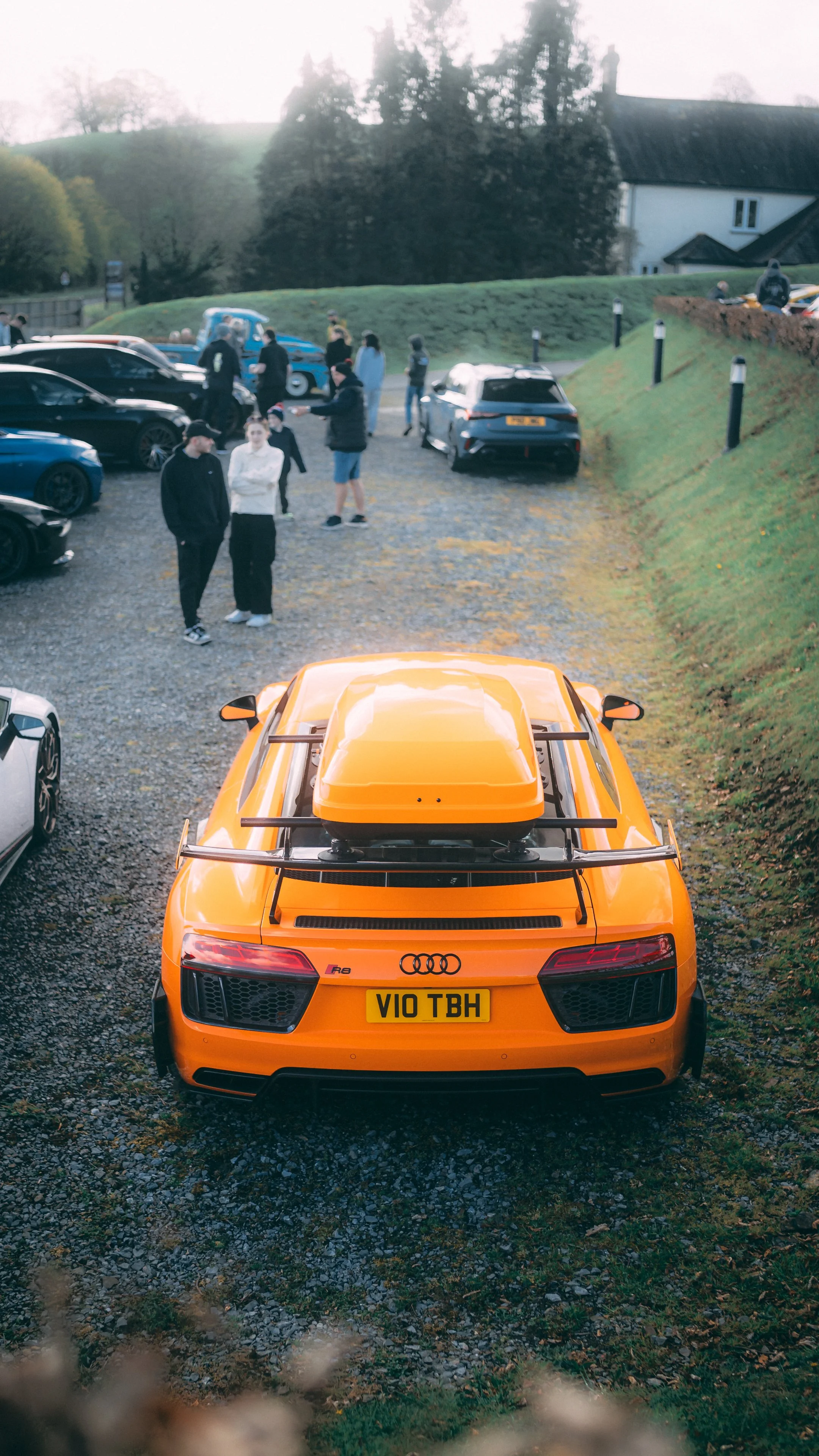 Orange Audi R8 sports car parked on gravel with a group of people and other cars in the background.