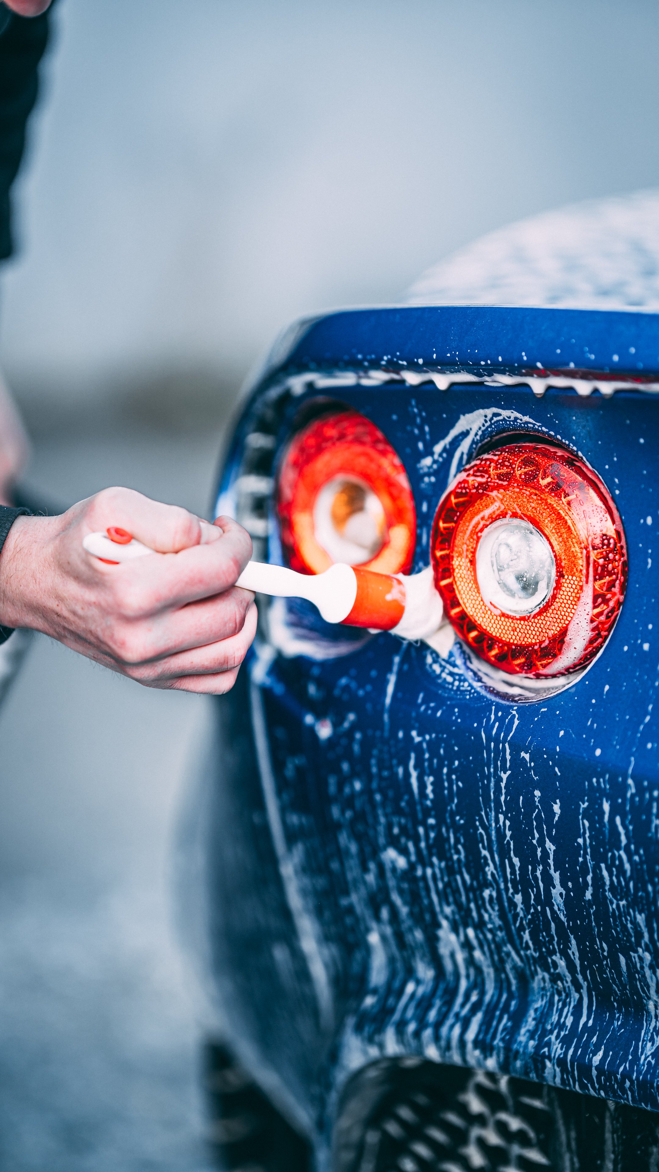 Close-up of a person applying soap to the taillights of a blue car during a wash.
