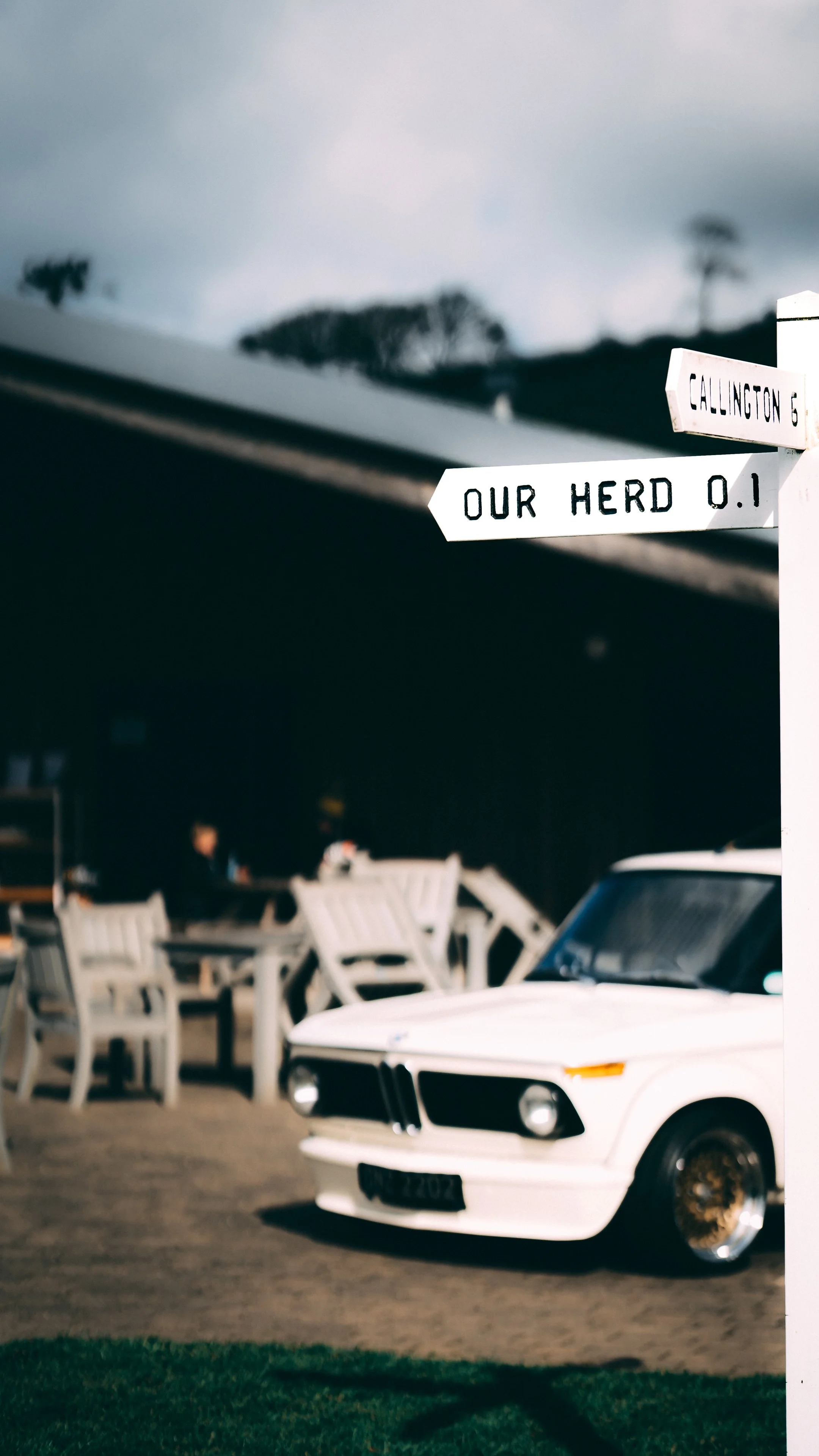 A white vintage car parked with outdoor seating and a signpost that reads "Gallington" and "Our Herd 0.1" in the background.