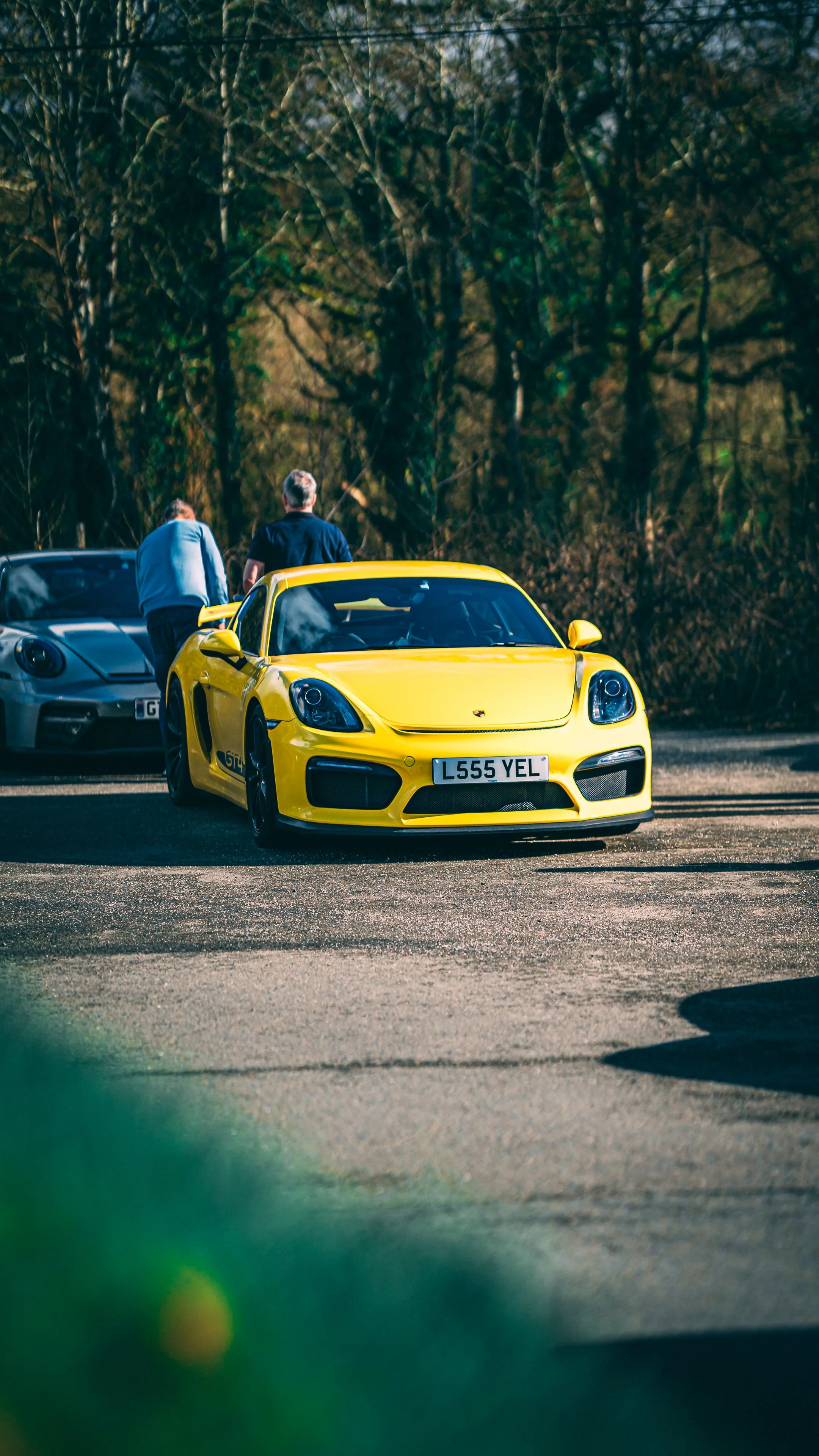 A yellow sports car with a UK license plate parked outdoors, with two men standing nearby and a forested background.