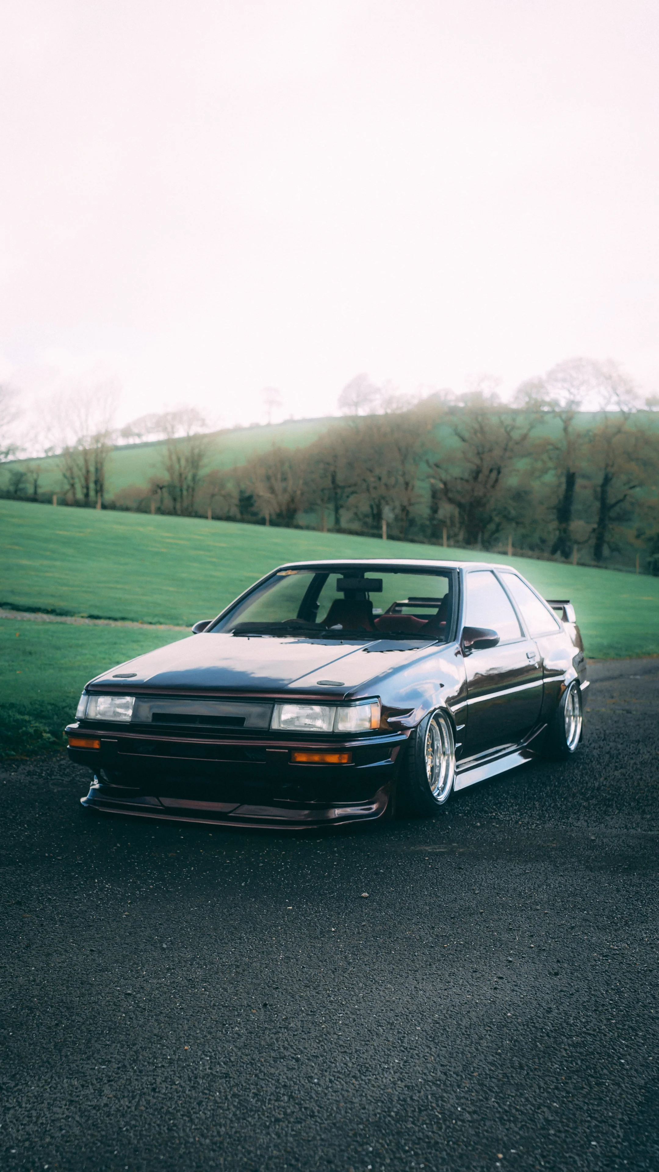 A black vintage sports car parked on a paved road with a grassy hill and trees in the background.