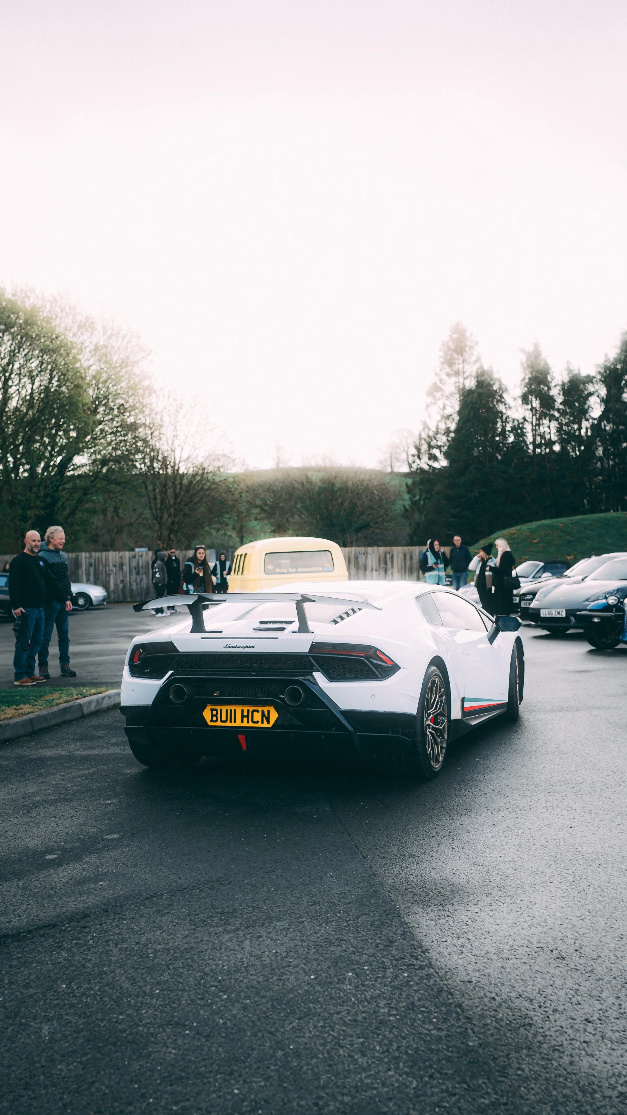 White Lamborghini sports car with a black rear spoiler and custom UK license plate BUII HCN parked in a lot during daytime, surrounded by other cars and people.