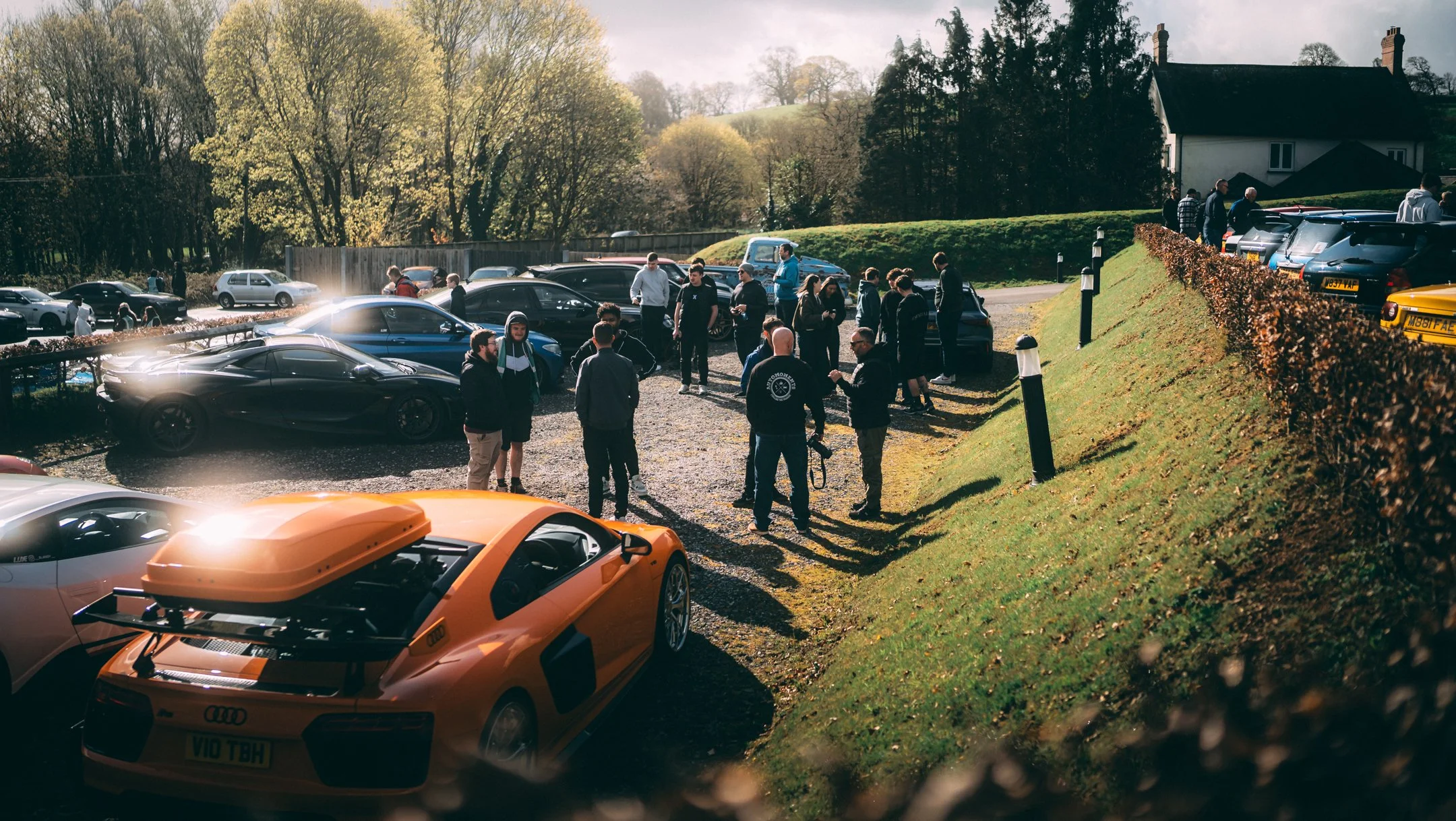 A gathering of people around parked cars in an outdoor area during daylight, with trees and a house in the background.