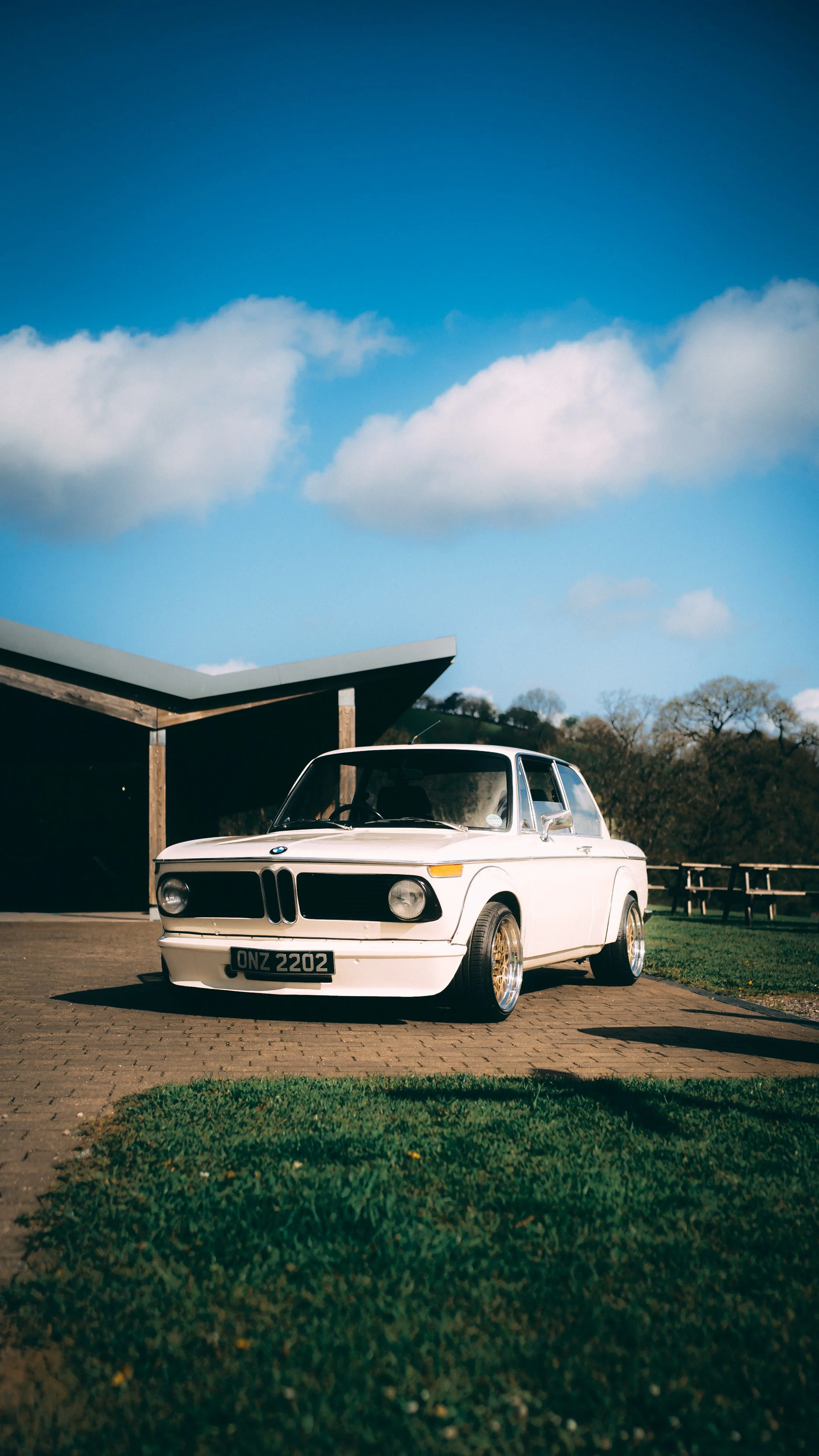 A white vintage BMW car parked on a brick driveway with a blue sky and clouds in the background.