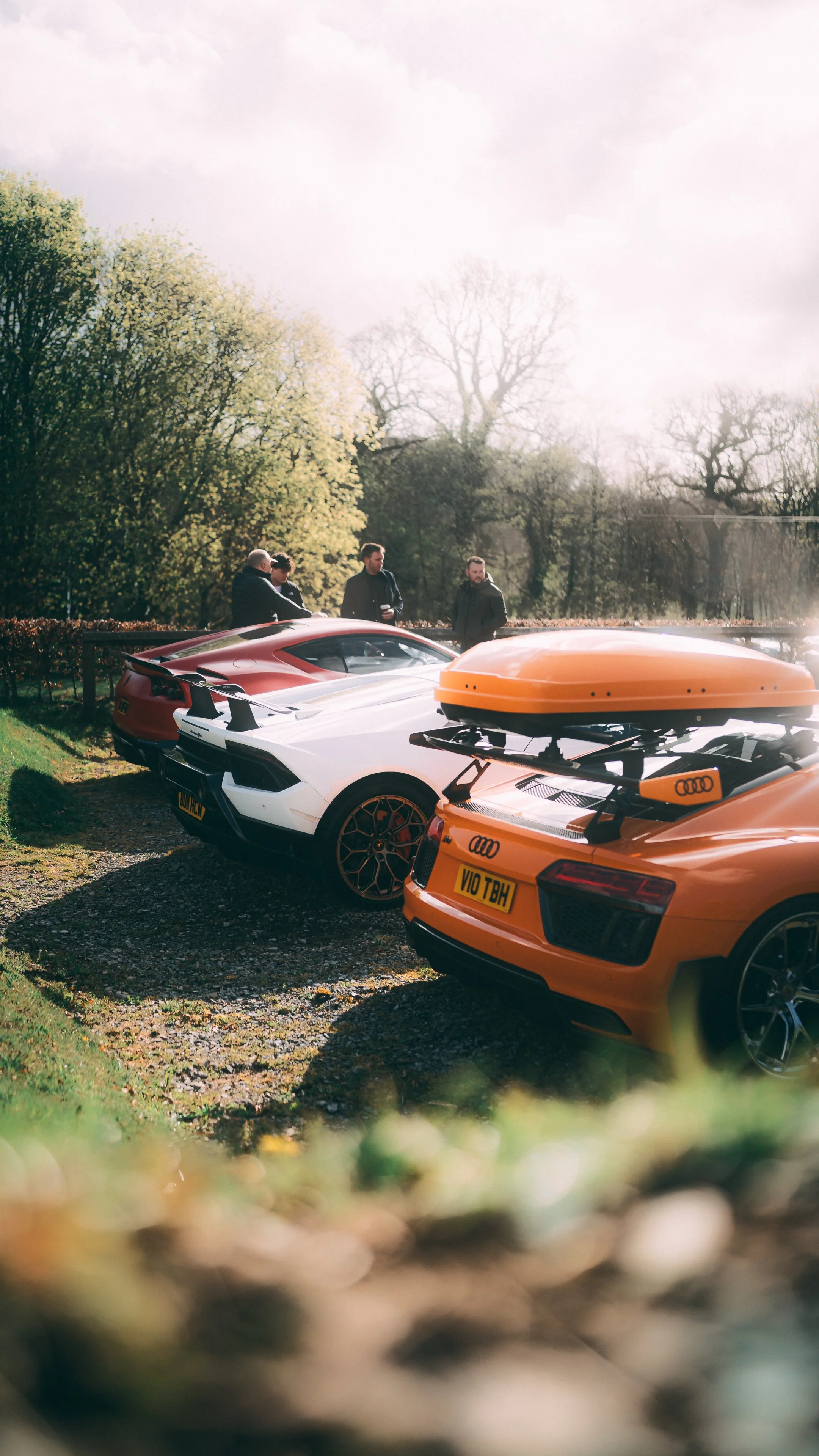 Four high-end sports cars parked outdoors in a row, with three men standing nearby and trees in the background.