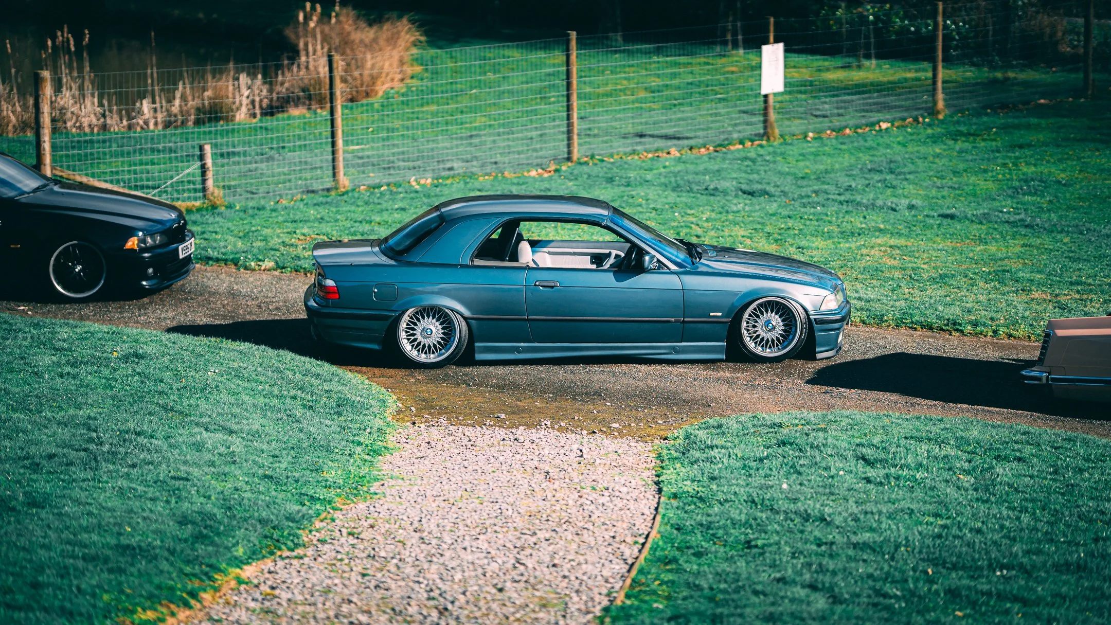 Three cars parked on a gravel driveway next to a grassy field with a wire fence in the background