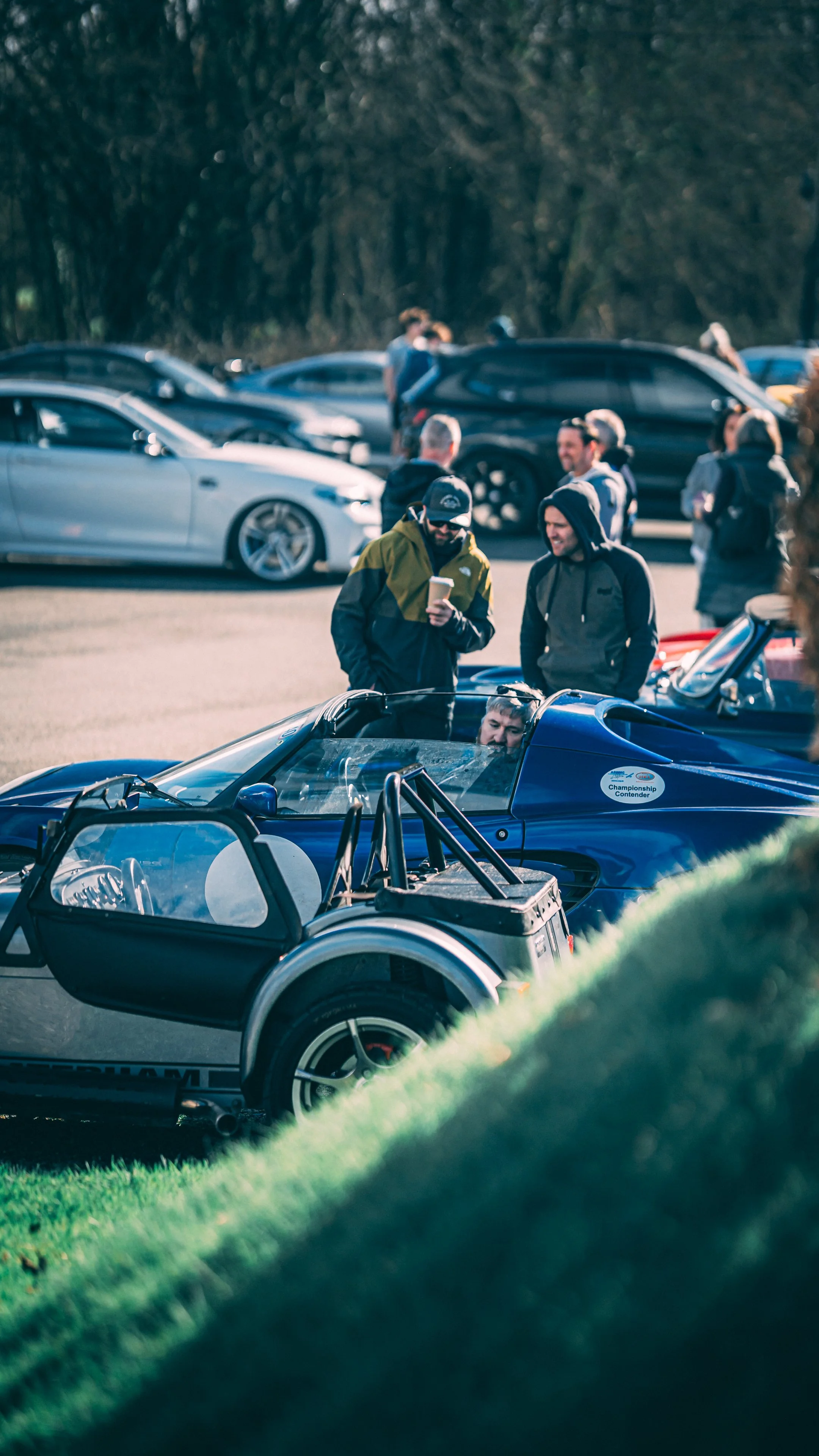 People gathered around a racing car at an outdoor event, with various cars parked in the background.