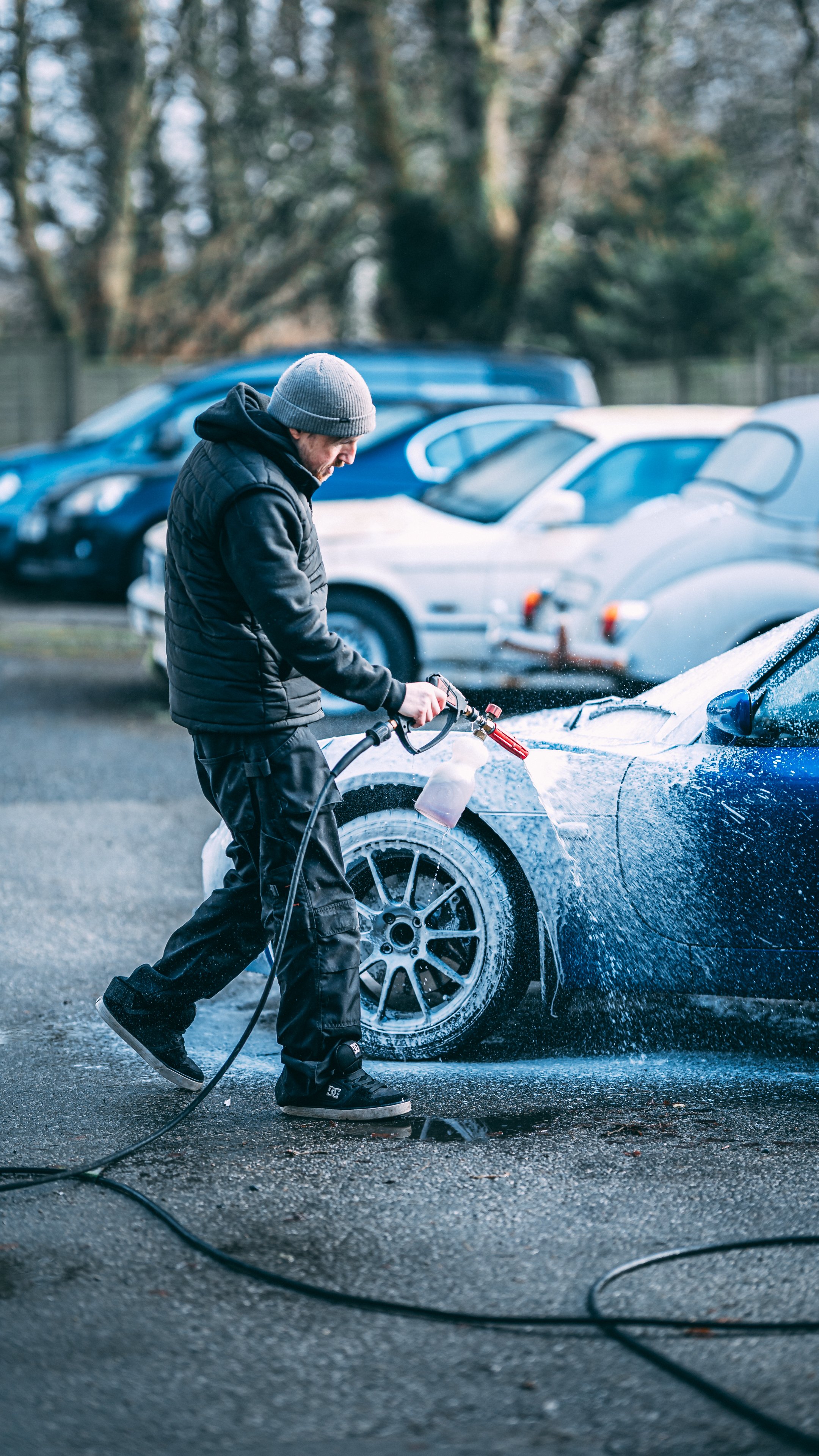 Man washing a blue sports car with a foamy soap during daytime in a parking lot.