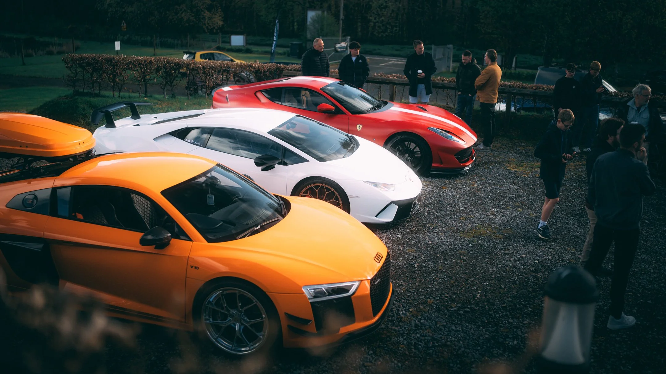 A group of people standing near a parking lot with several sports cars, including an orange Audi, a white Lamborghini, and a red Ferrari.