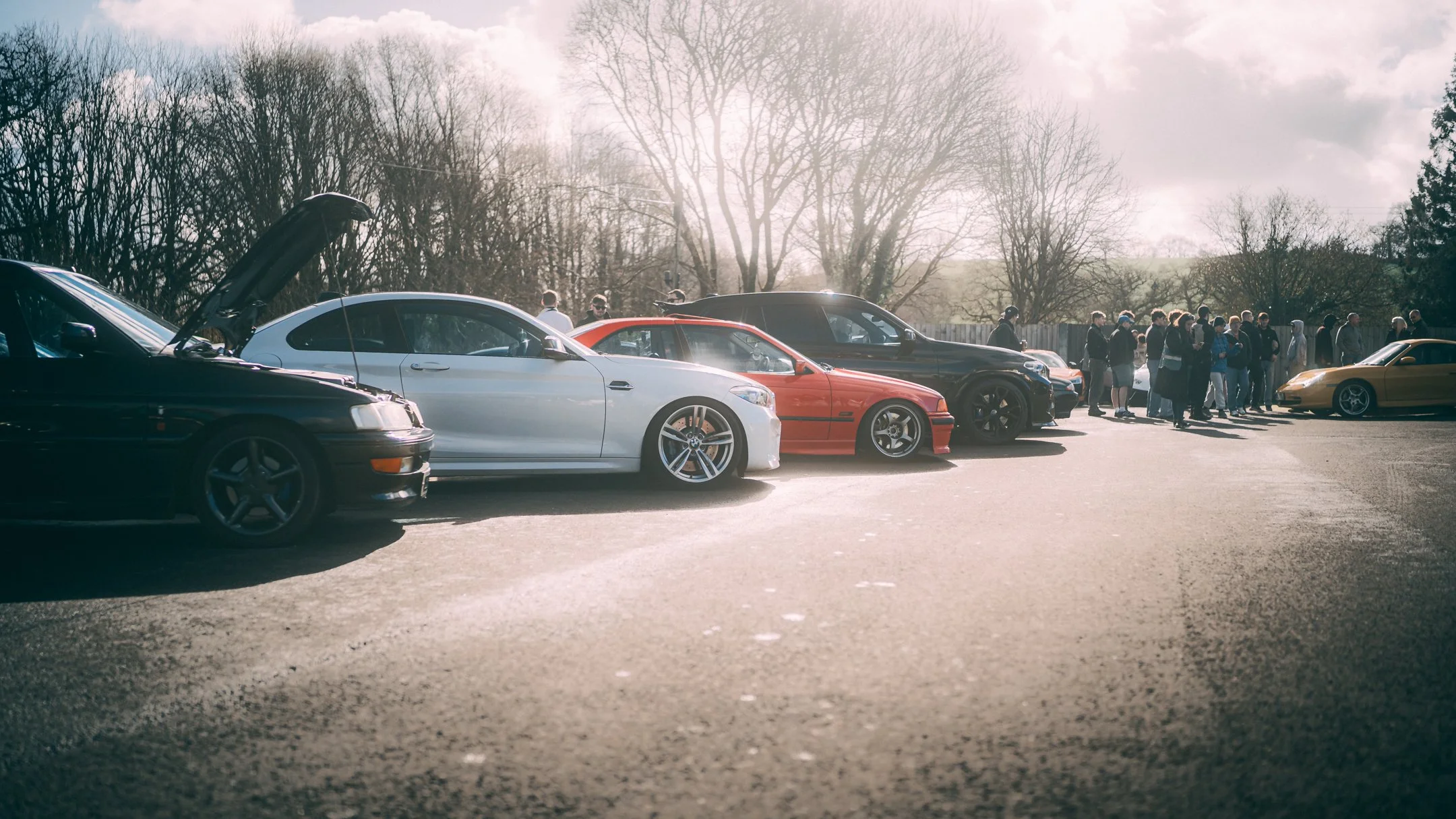 A row of sports cars parked in a lot with their hoods open and people standing nearby, against a backdrop of leafless trees and a partly cloudy sky.
