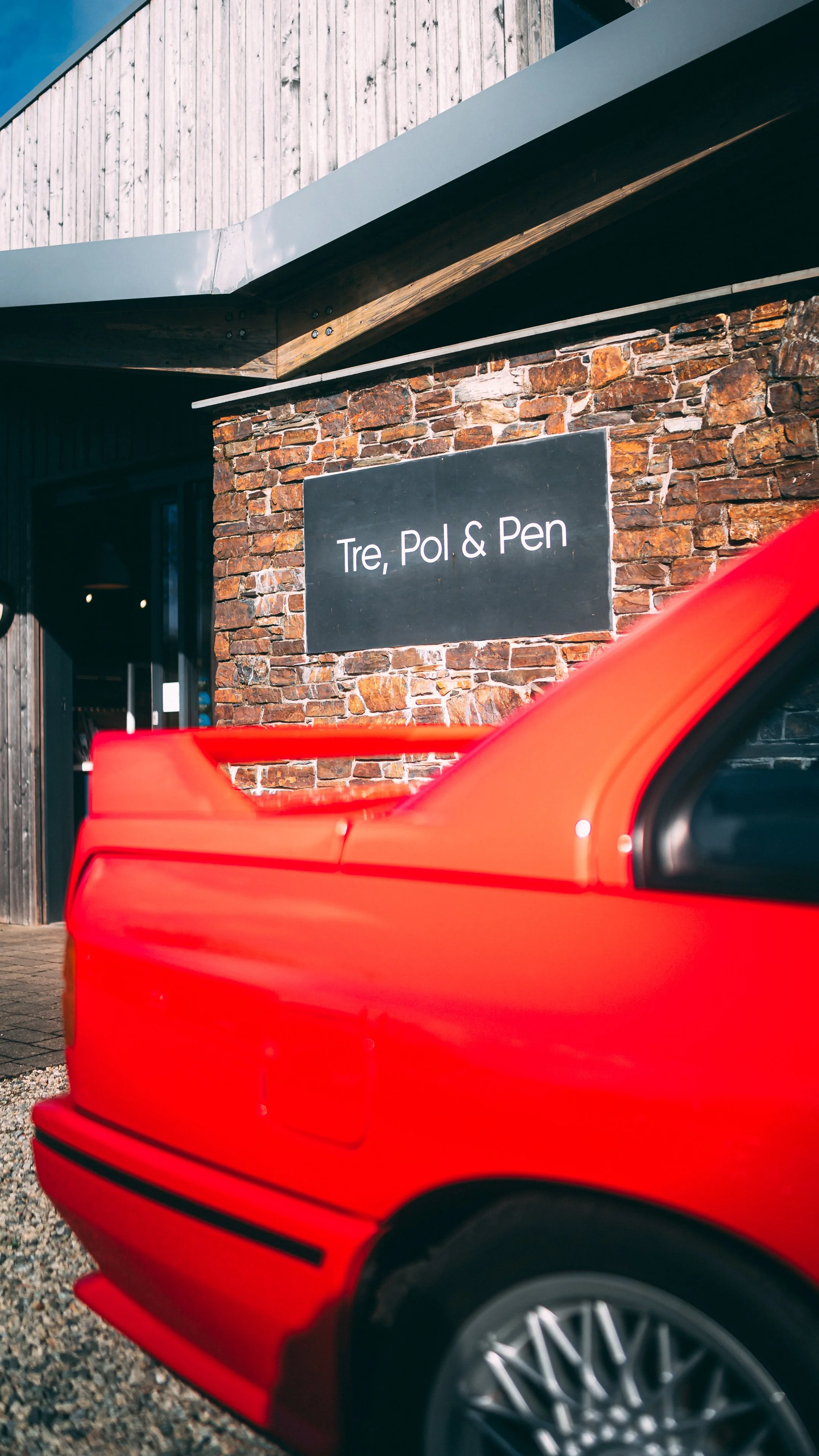 Part of a red sports car with a black wheel, parked in front of a building with a brick wall and a sign that reads "Tre, Pol & Pen".