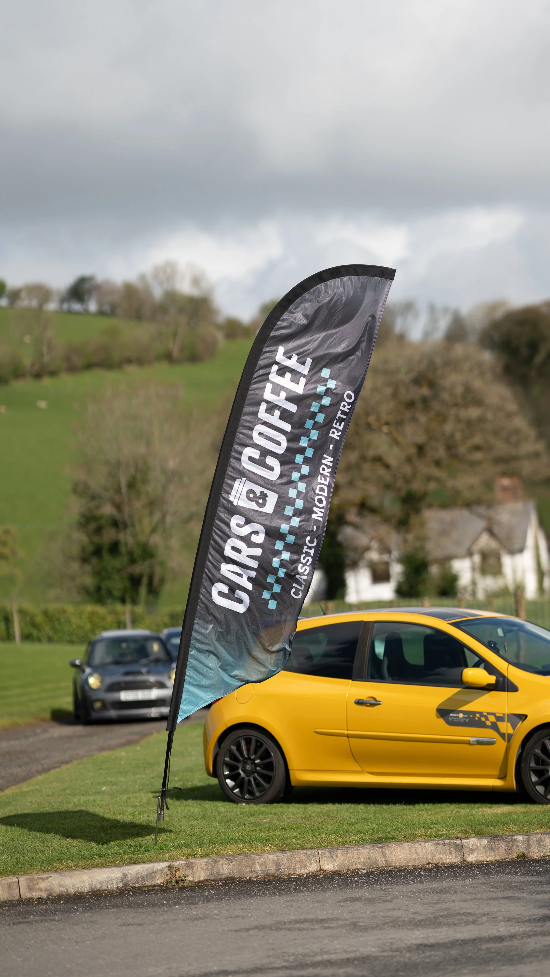 A black and blue flag that reads "Cars & Coffee Classic, Modern, Retro" is positioned on a grassy area near parked cars, including a yellow car and a black car, with a cloudy sky and green hills in the background.