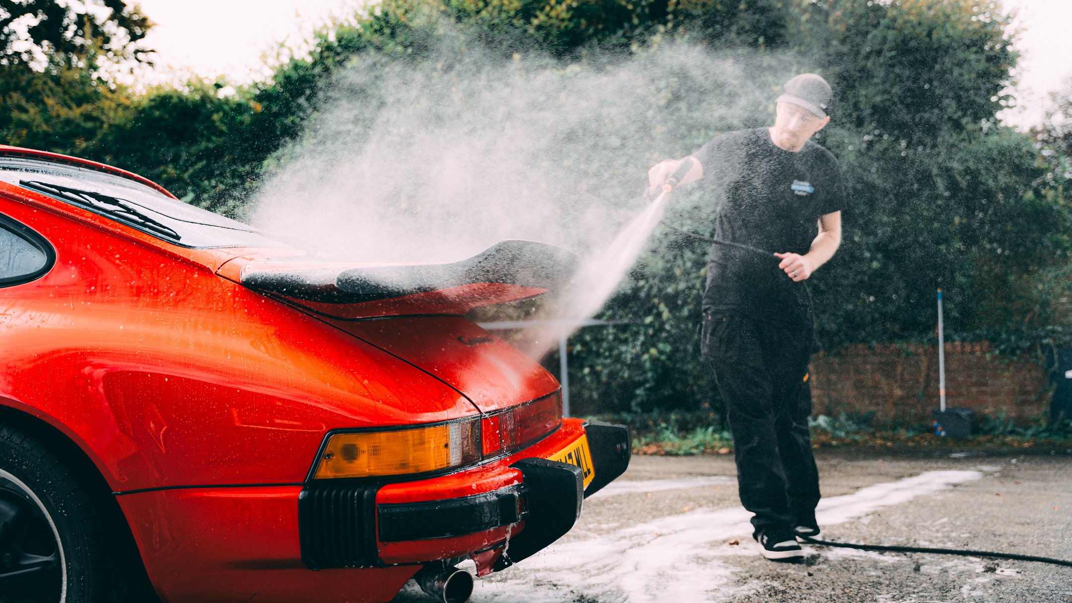 Person washing a red sports car outdoors with a garden in the background.