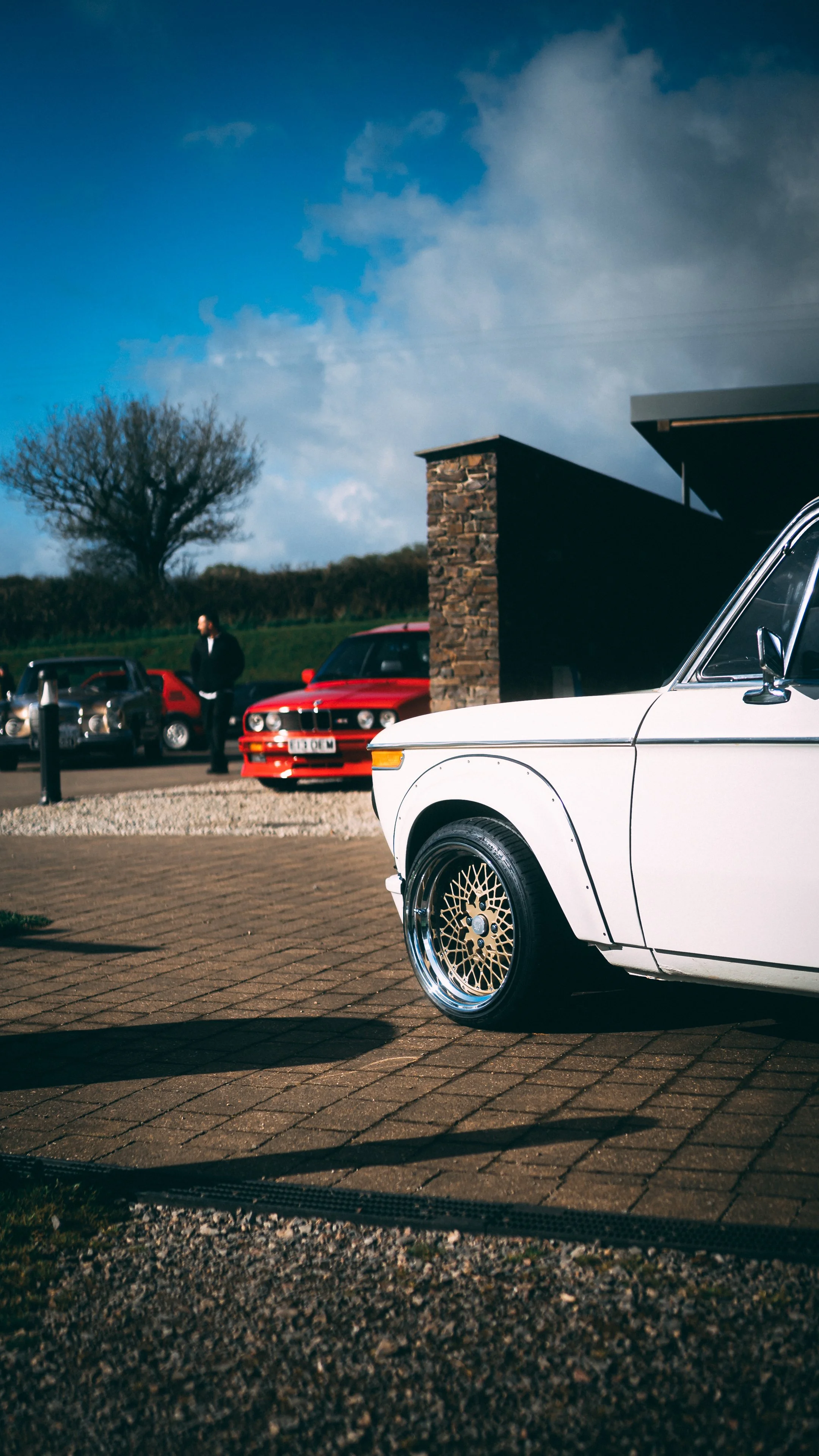 Close-up of a white vintage car with custom rims parked on a brick driveway, with other classic cars and a man in the background under a partly cloudy sky.